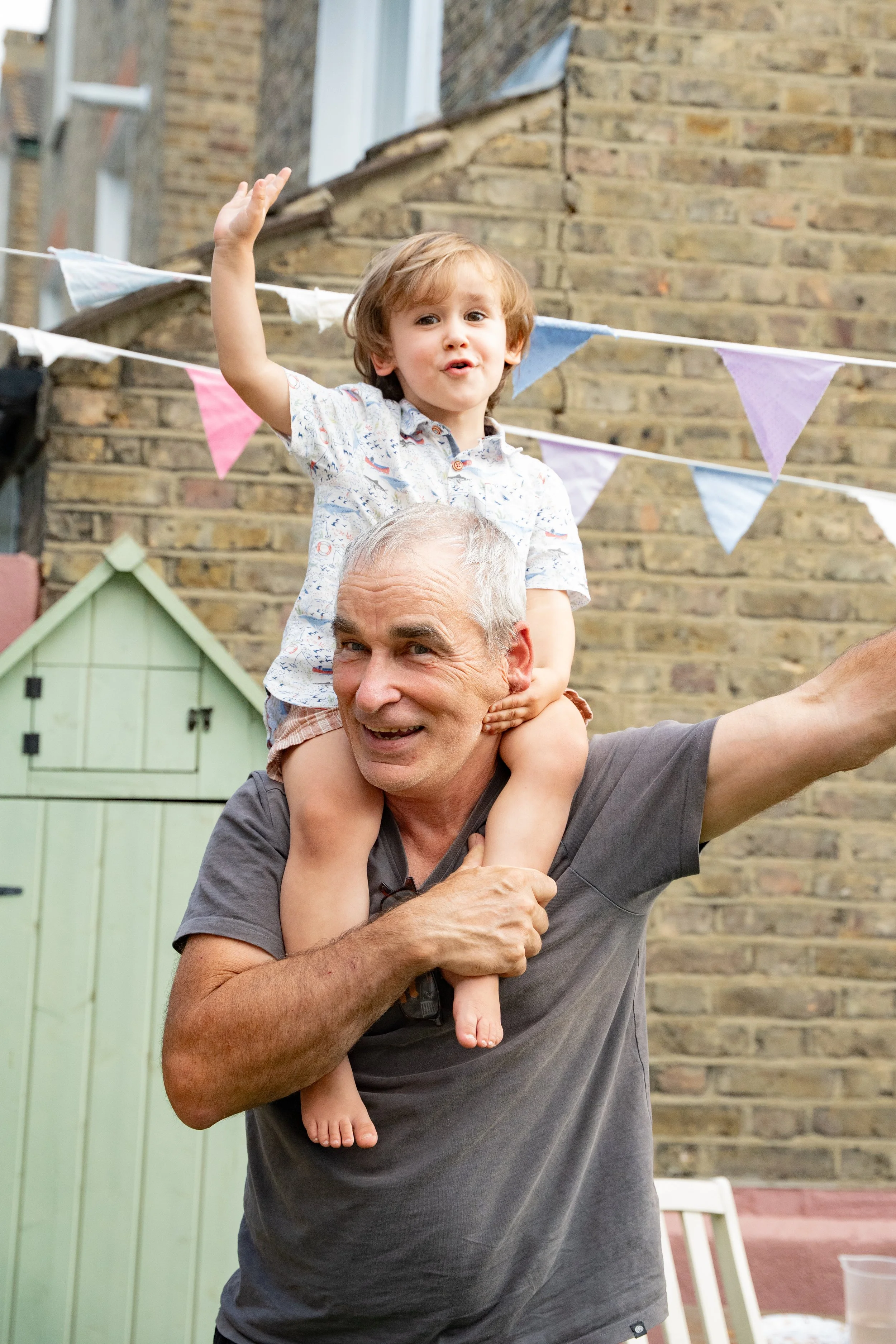 A happy elderly man with white hair carrying a young boy on his shoulders during an outdoor gathering. The boy is waving and smiling. Colorful bunting hangs in the background, with a brick building behind them.