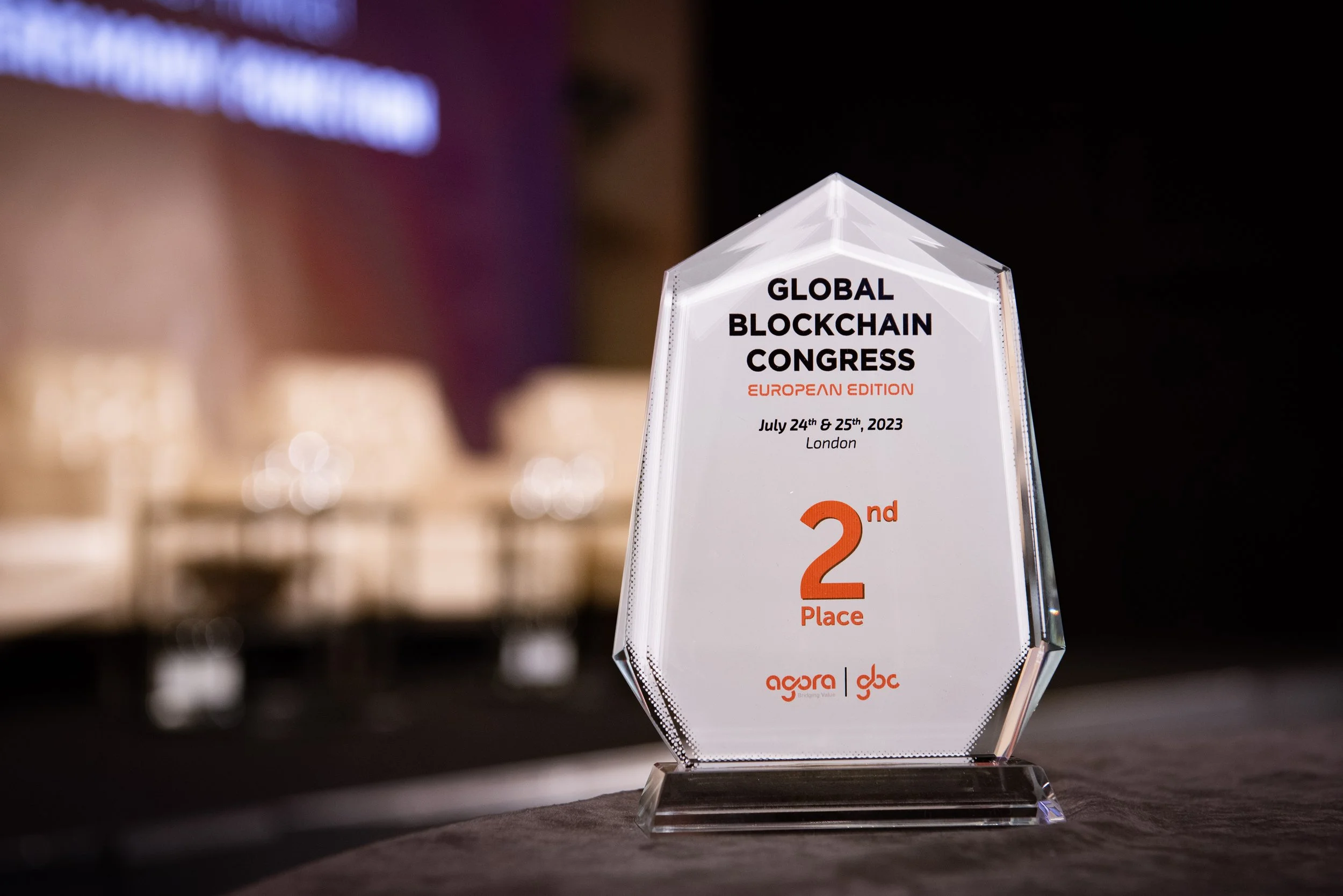 A clear glass award with beveled edges on a black base, inscribed with details of the Global Blockchain Congress European Edition, held July 24-25, 2023, in London, indicating second place. The background is blurred with warm lighting.