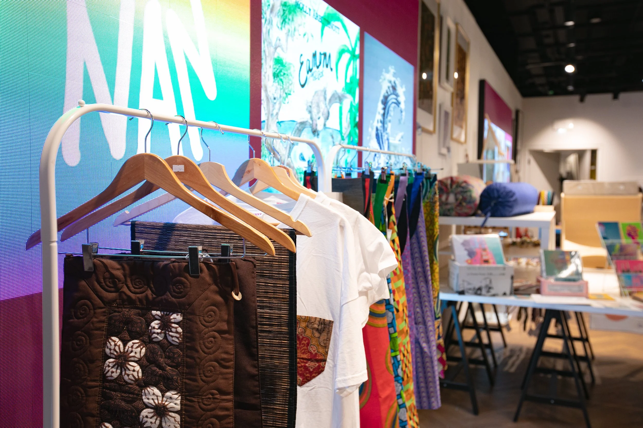 Clothing rack displaying various garments including patterned skirts and white T-shirts, with colorful framed artwork on the wall and tables with books and merchandise in an indoor retail space.