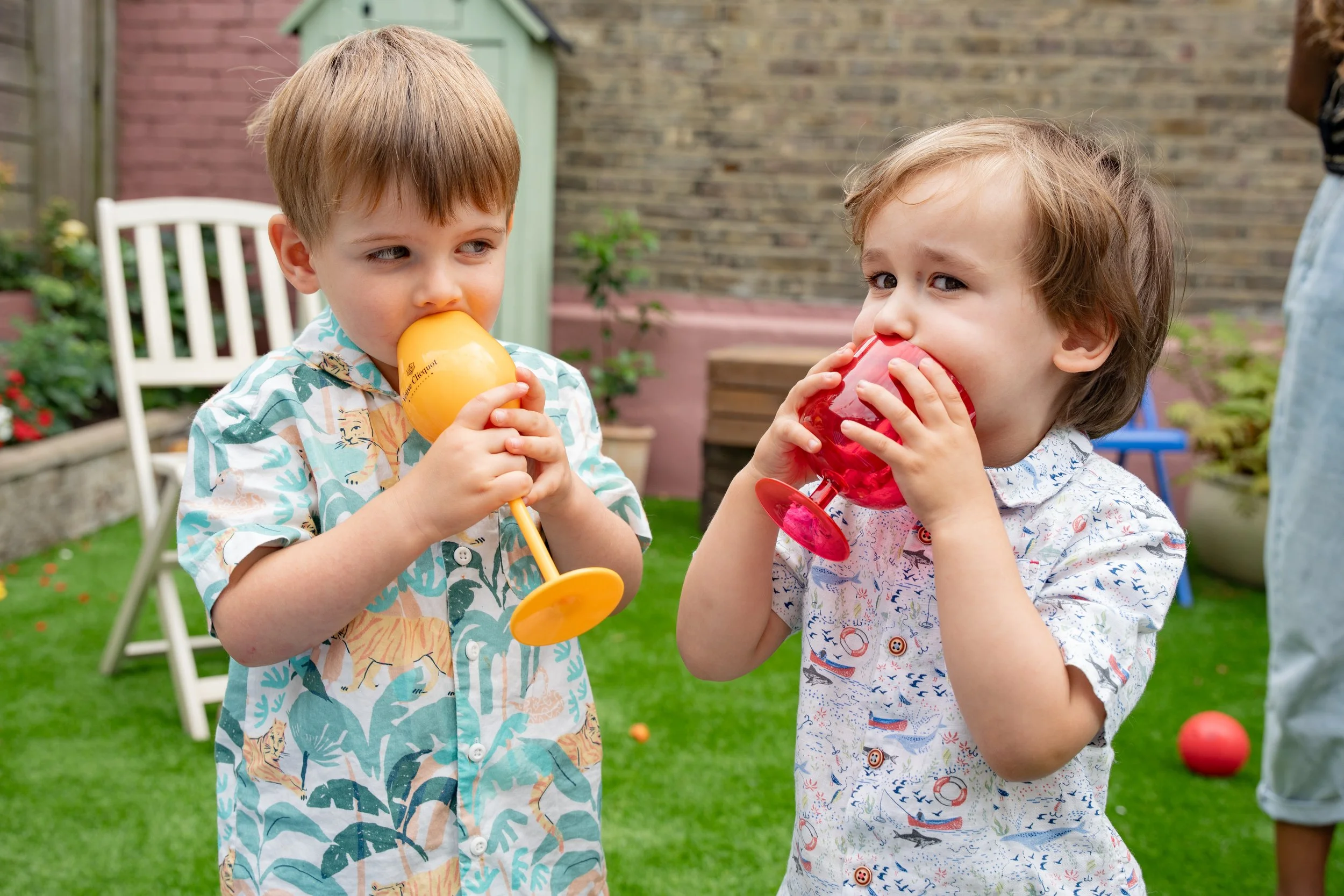 Two young boys with drinks in colorful plastic wine glasses at an outdoor gathering on a lawn, with garden furniture and a brick wall in the background.