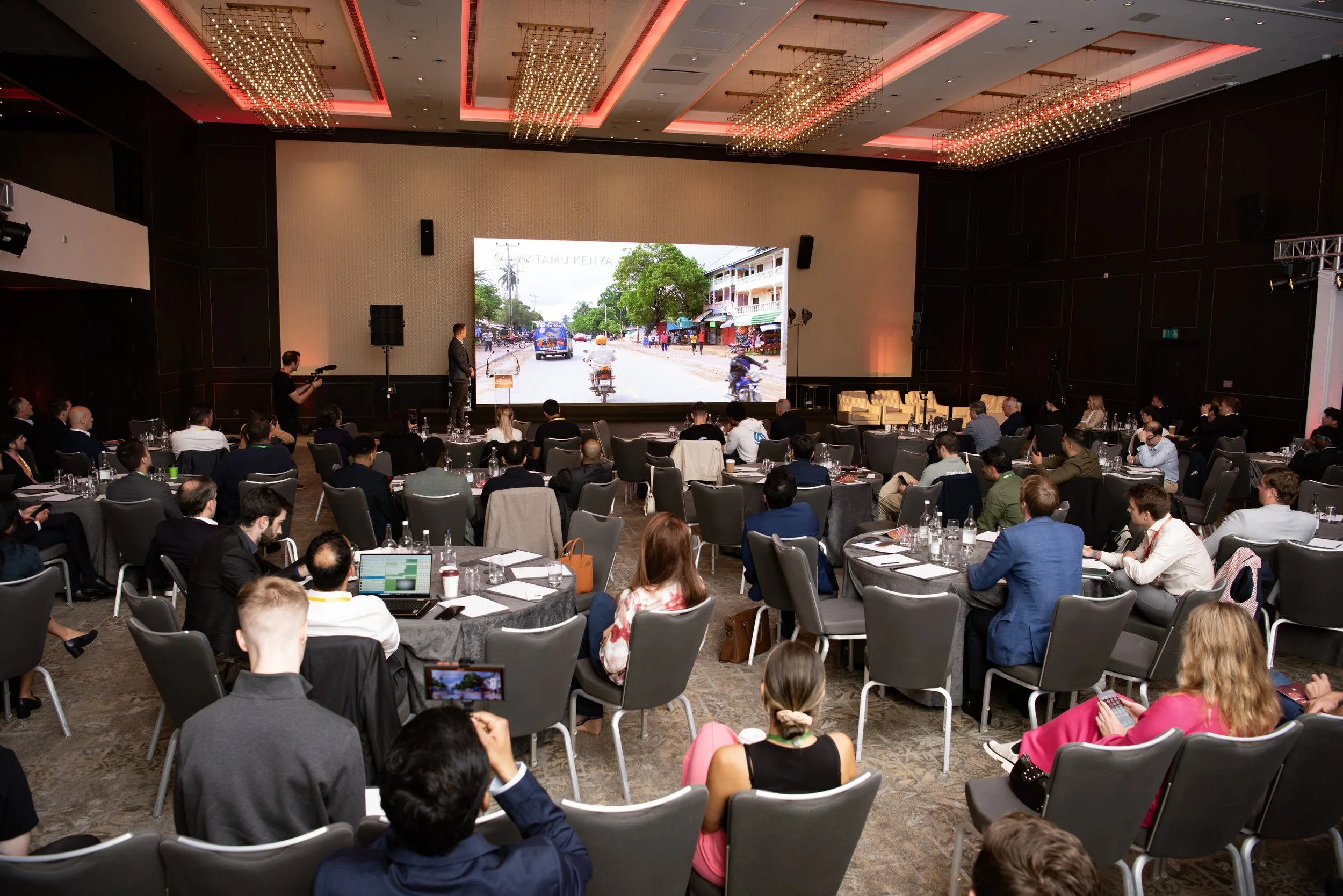 Attendees seated at round tables in a conference room watching a presentation on a large screen showing a street scene in Kenya.