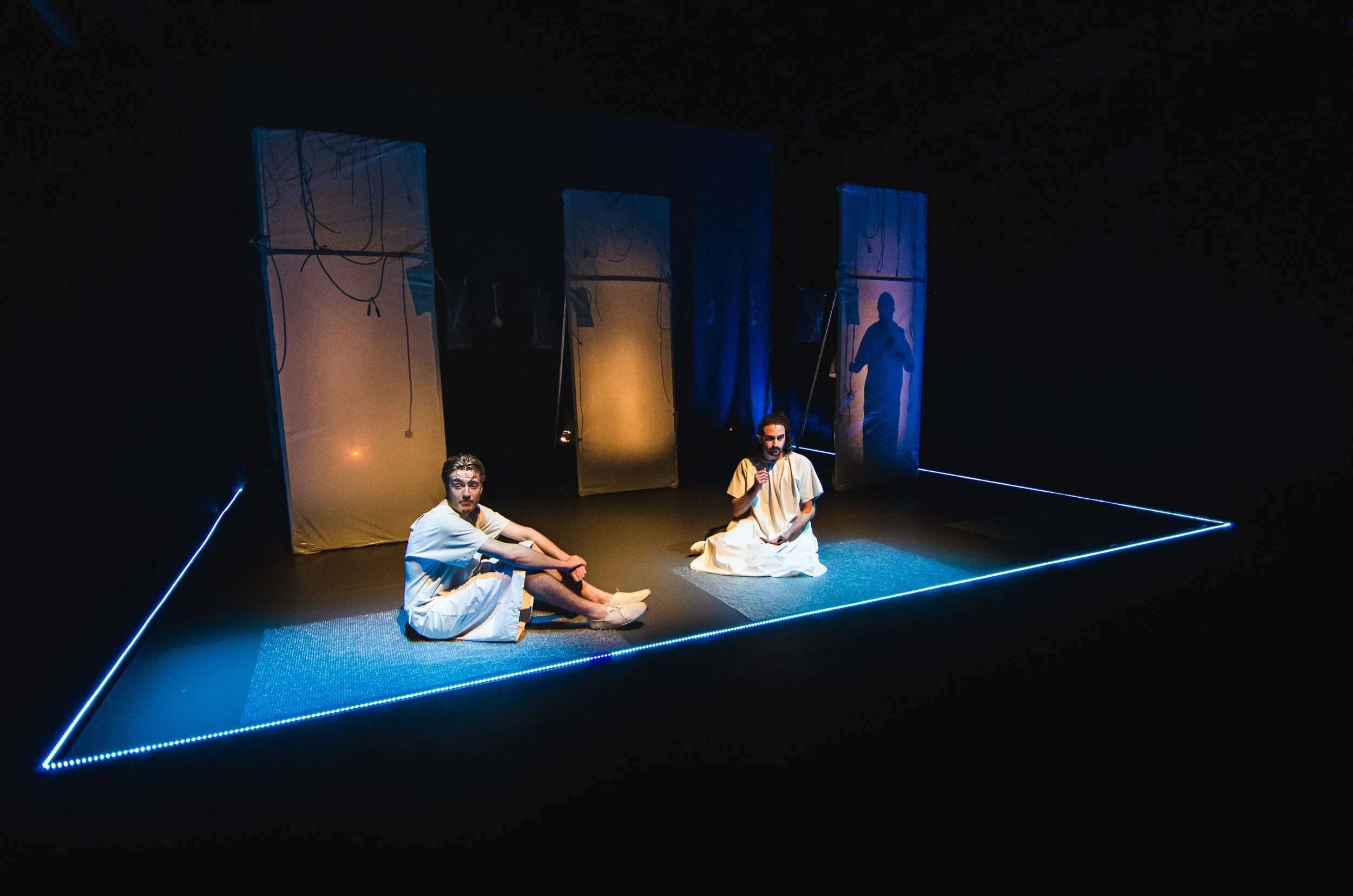 Two performers dressed in white robes sitting on stage with blue lighting and semi-transparent hanging screens in the background, creating a theatrical atmosphere.