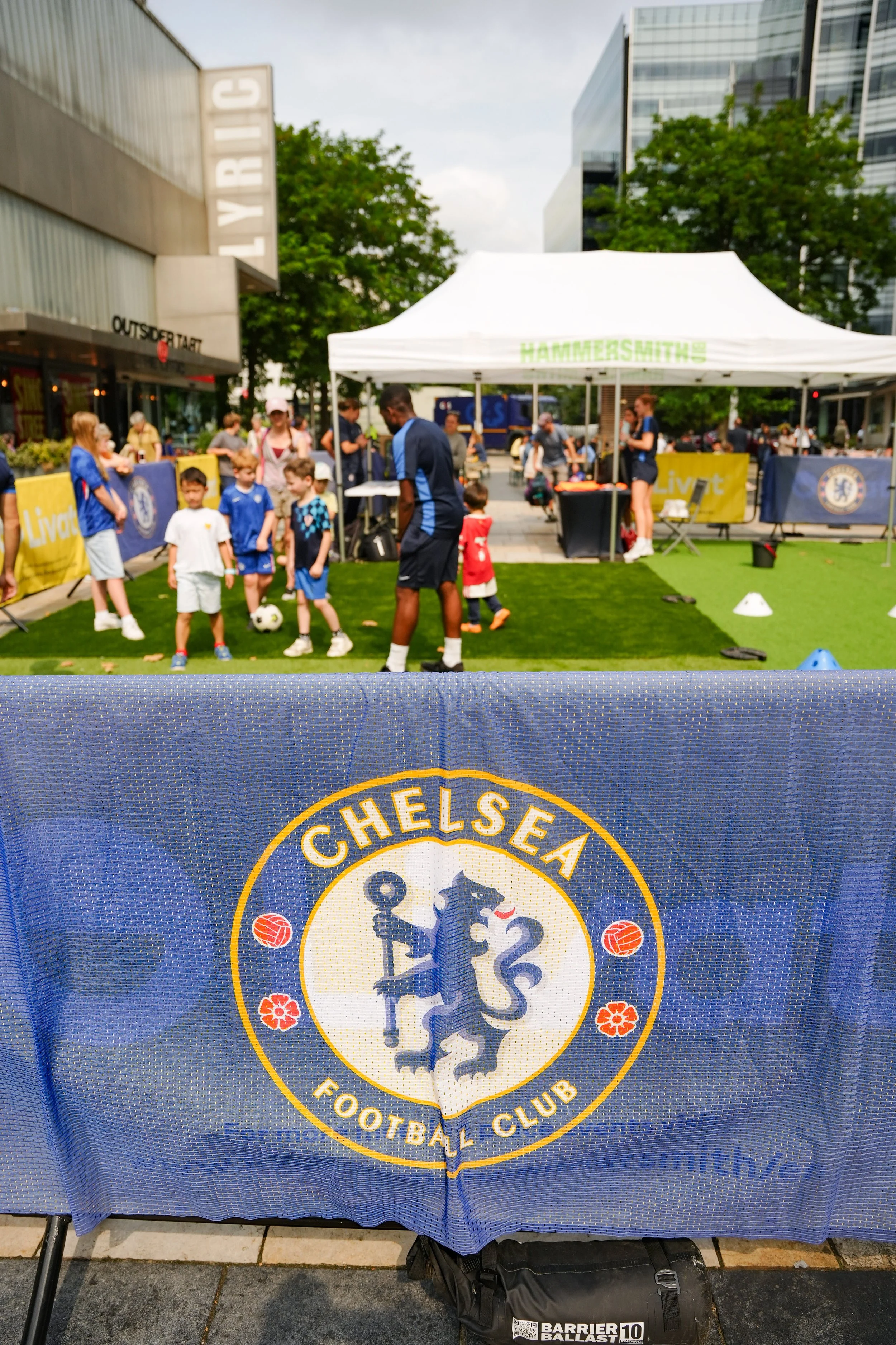 Chelsea Football Club banner in front of a public event where children are playing and adults are supervising under a white tent with a sign that reads 'HAMMERSMITHS', with buildings and trees in the background.