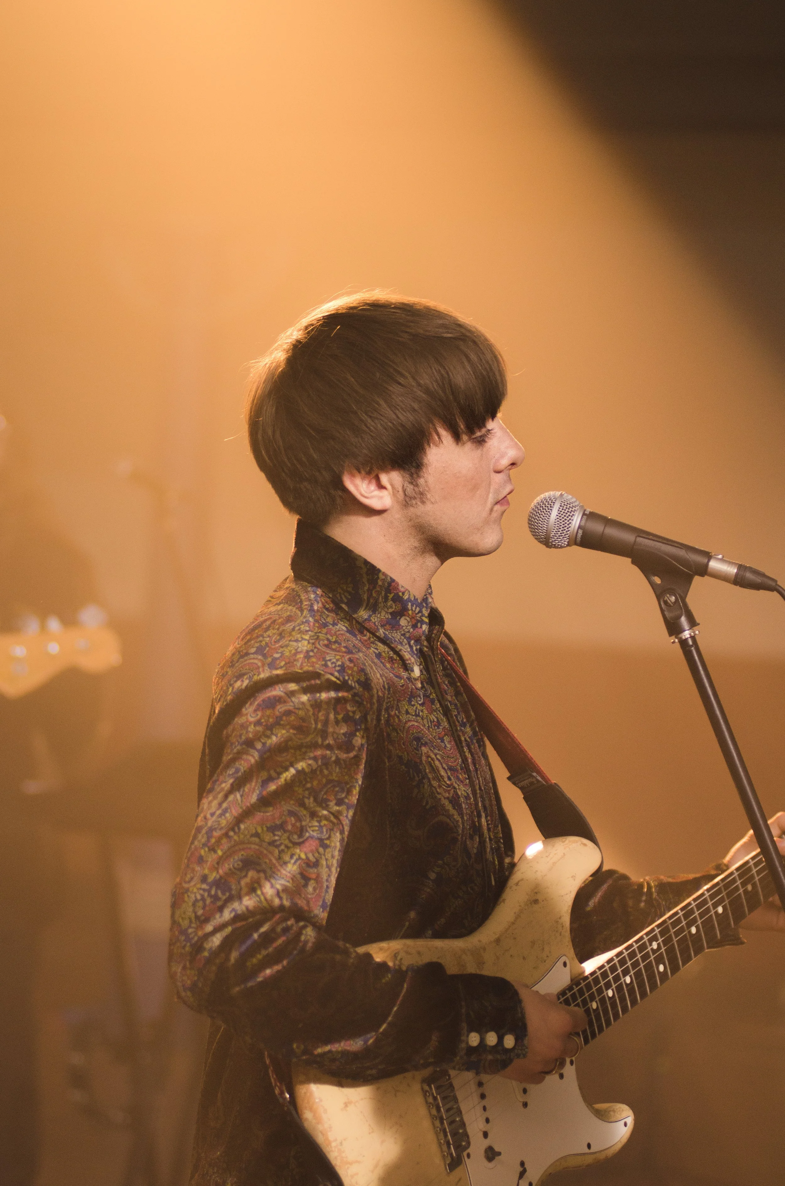 Young man with dark brown hair playing an electric guitar on stage, wearing a colorful patterned jacket, with a microphone in front of him and warm stage lighting.