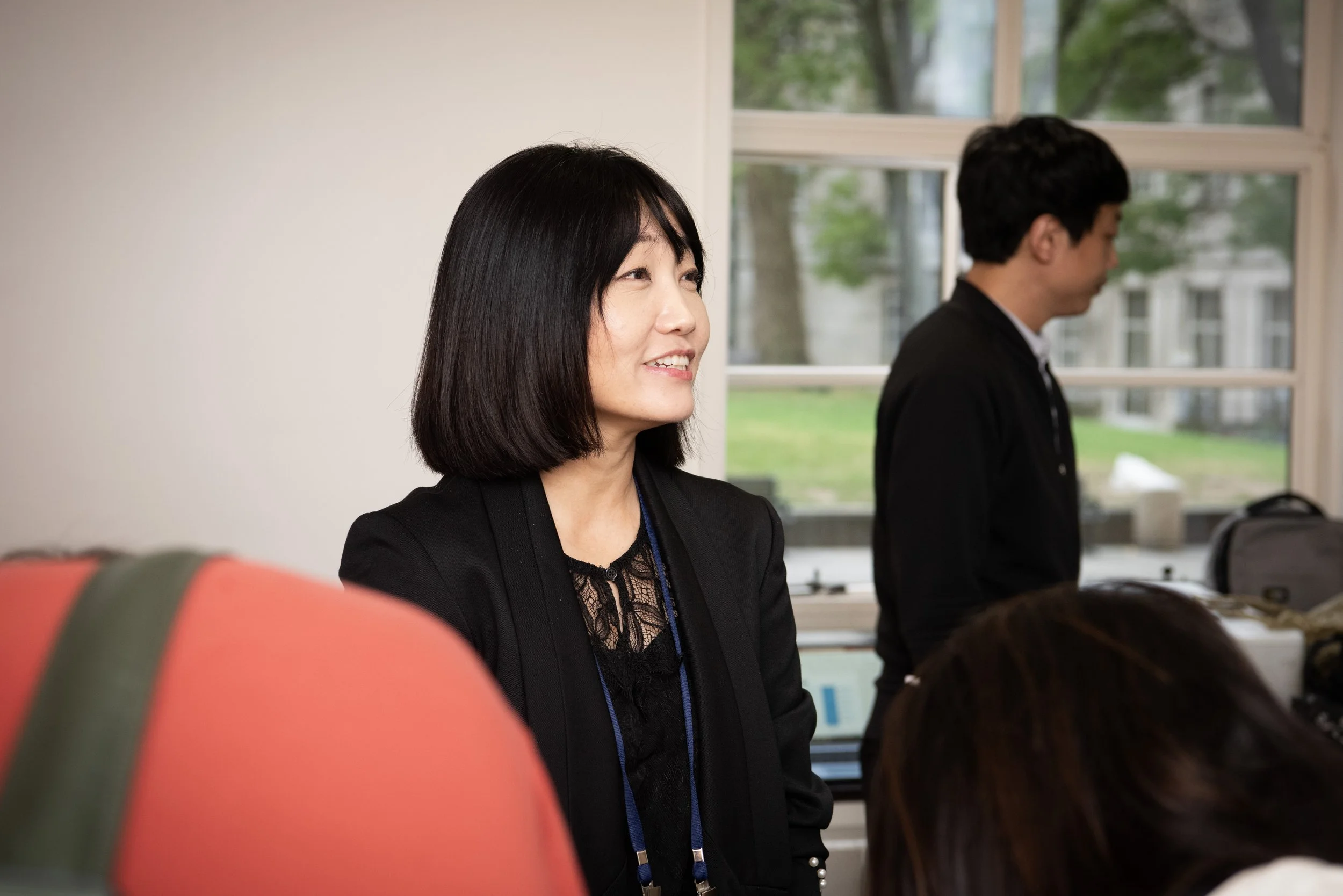 A woman with short black hair, wearing a black blazer and a lace top, smiling during a meeting or discussion in an office setting, with a man in a black blazer and white shirt in the background.