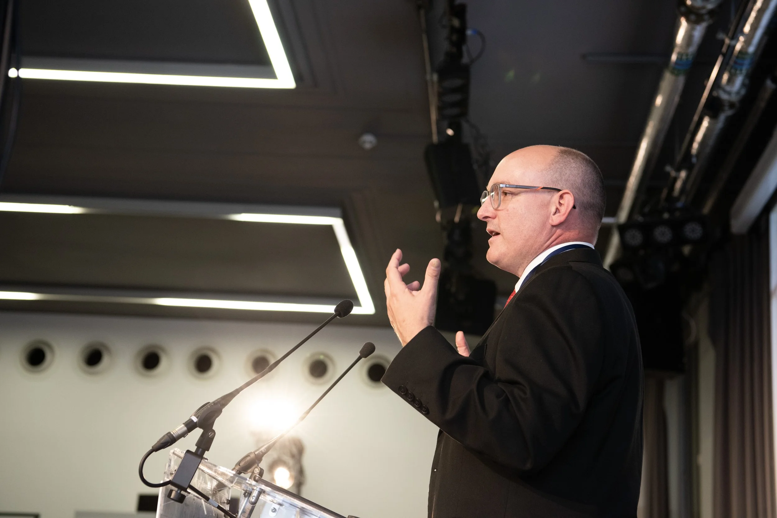 Side profile of a man in glasses and a suit speaking at a podium with microphones, indoors with modern ceiling light fixtures and stage equipment in the background.
