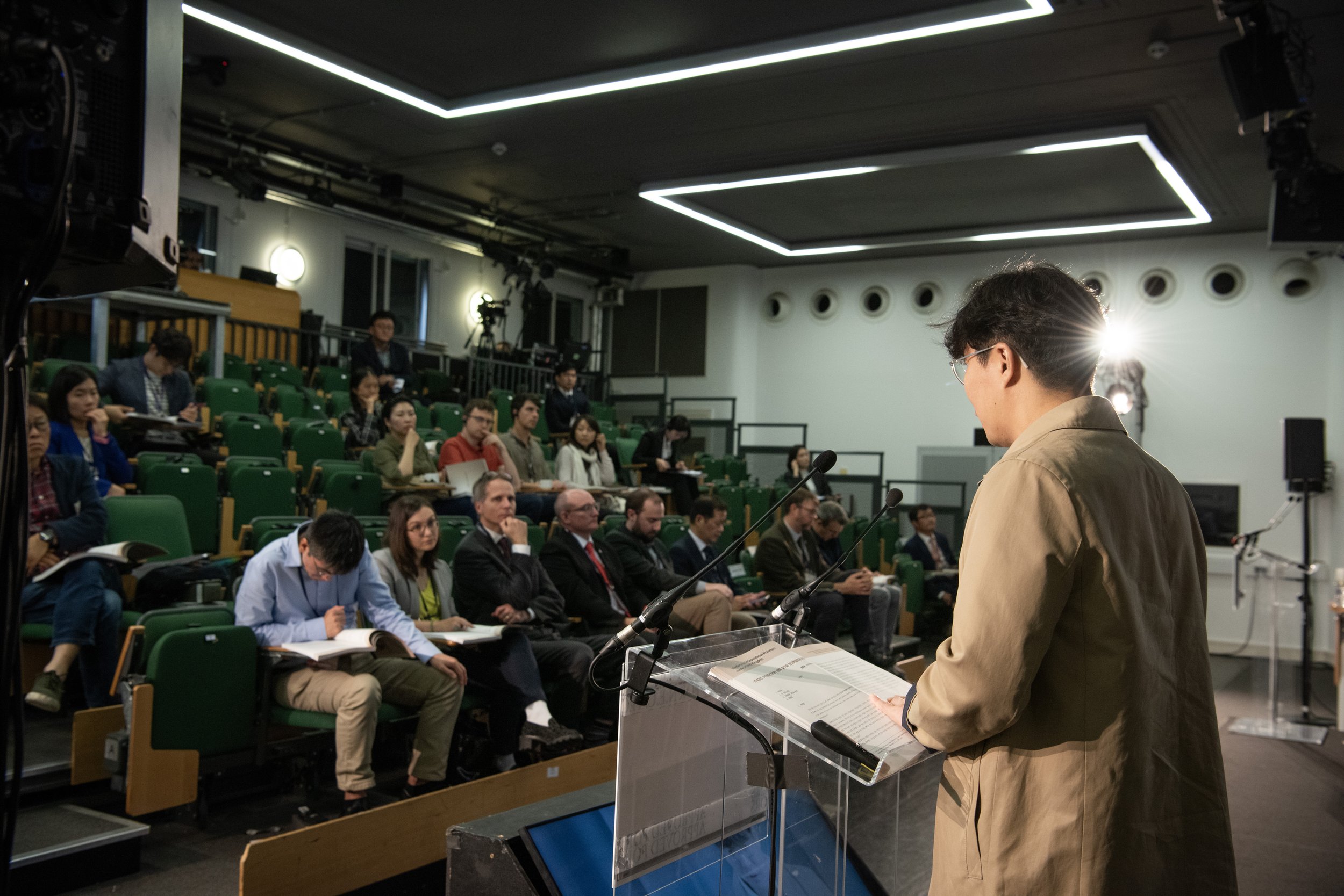 A man giving a speech at a conference or seminar, standing behind a clear podium with microphones, in front of an audience seated in green chairs in a lecture hall or conference room.