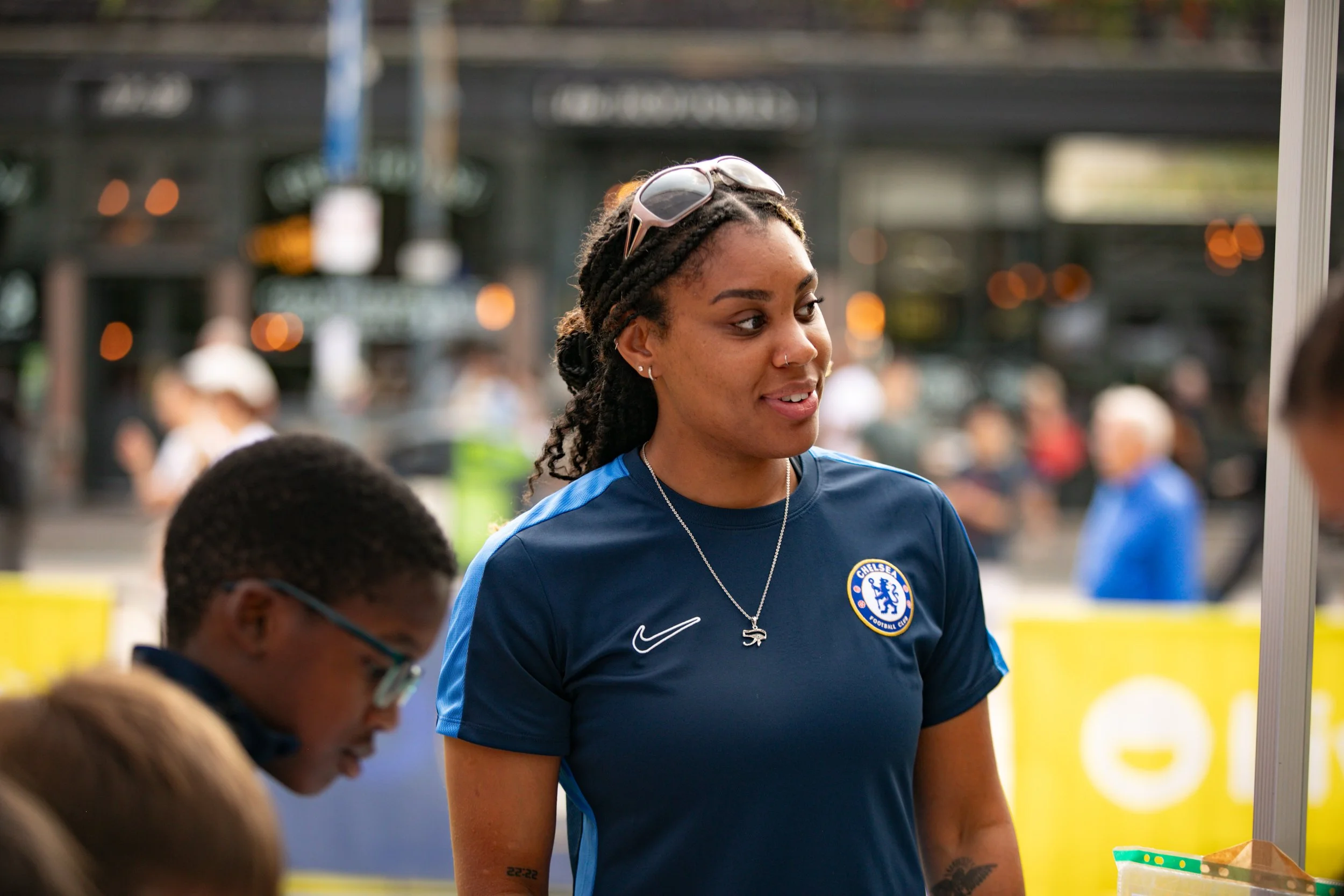 A young woman with dreadlocks, sunglasses on her head, wearing a Chelsea football club jersey, standing at an outdoor event, engaging in conversation with a young man.