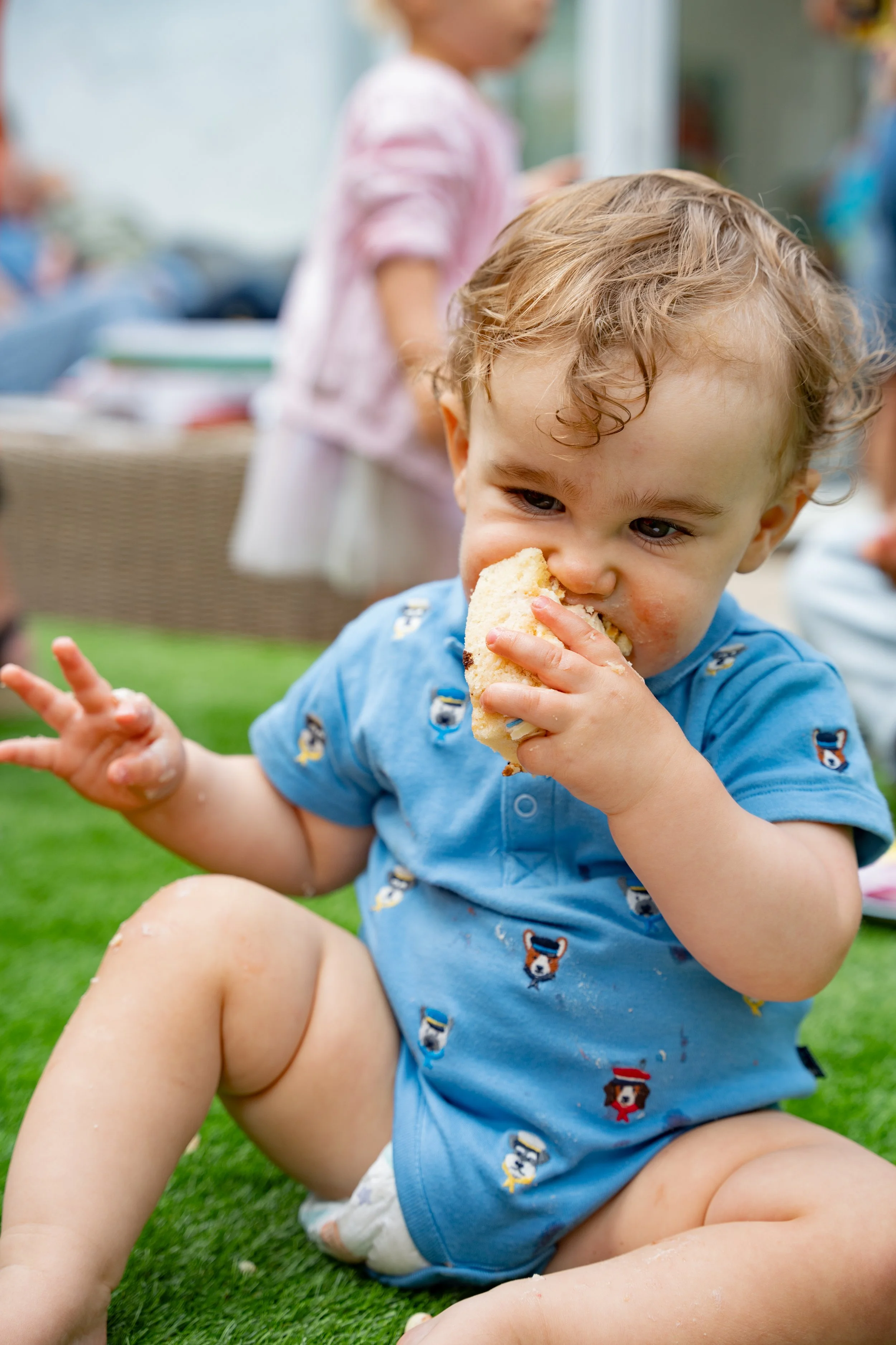 Young boy with curly blonde hair wearing a blue shirt with dog patterns, sitting on grass and eating a sandwich