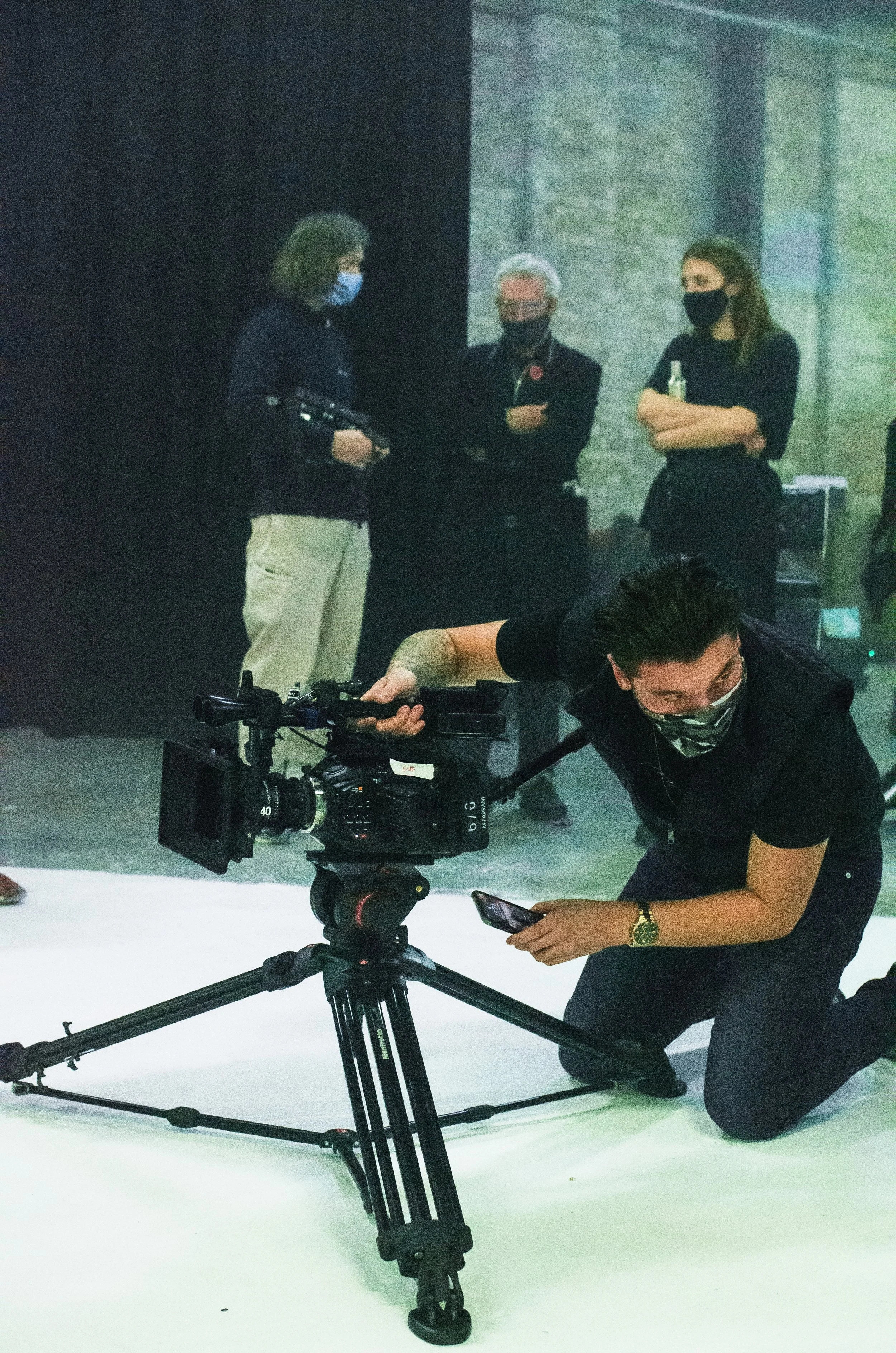 A person kneeling on the floor operating a professional video camera, with four people in the background wearing face masks and engaged in conversation.
