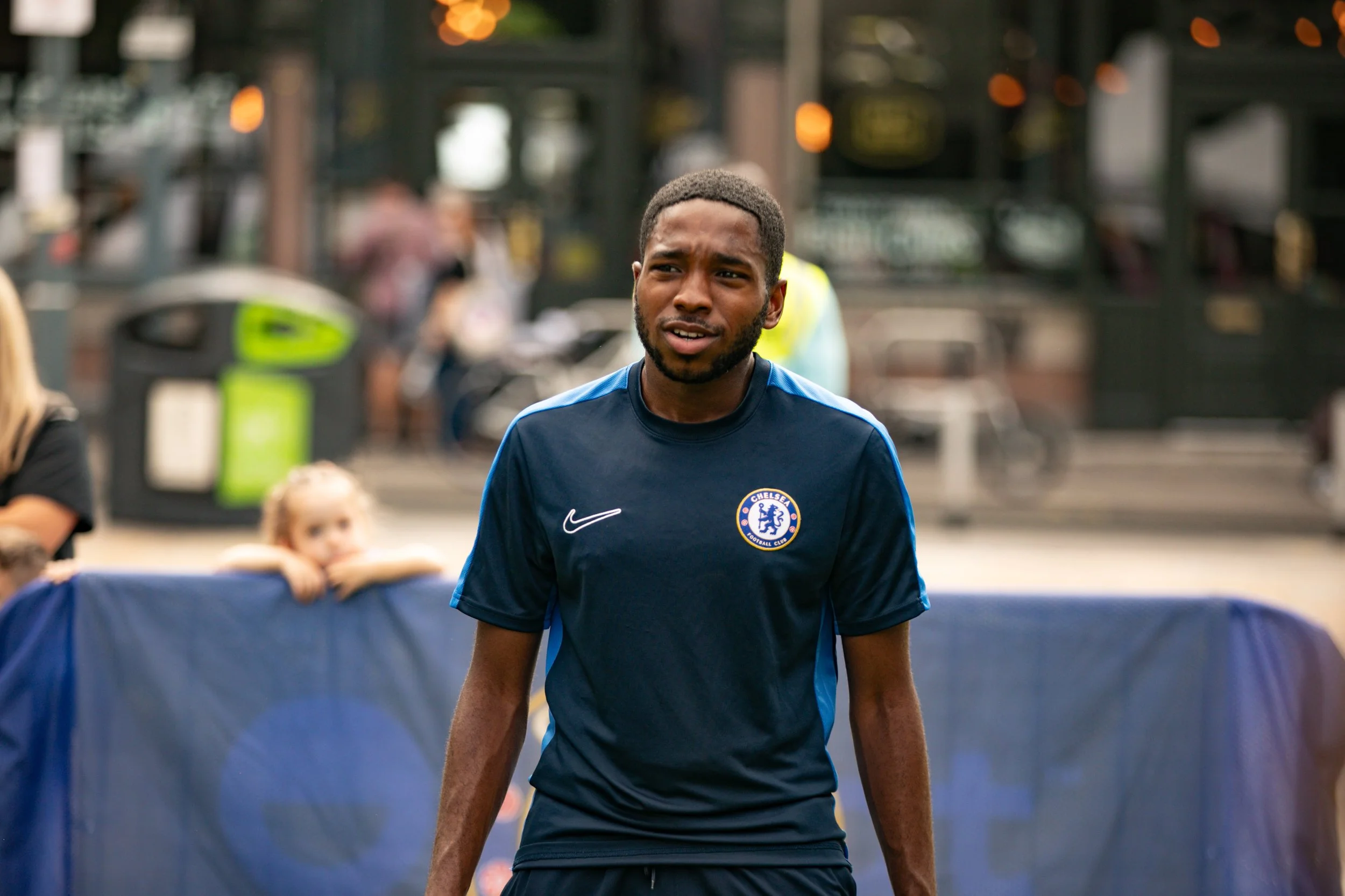 A young man in a blue Chelsea football jersey standing outdoors with a concentration expression.