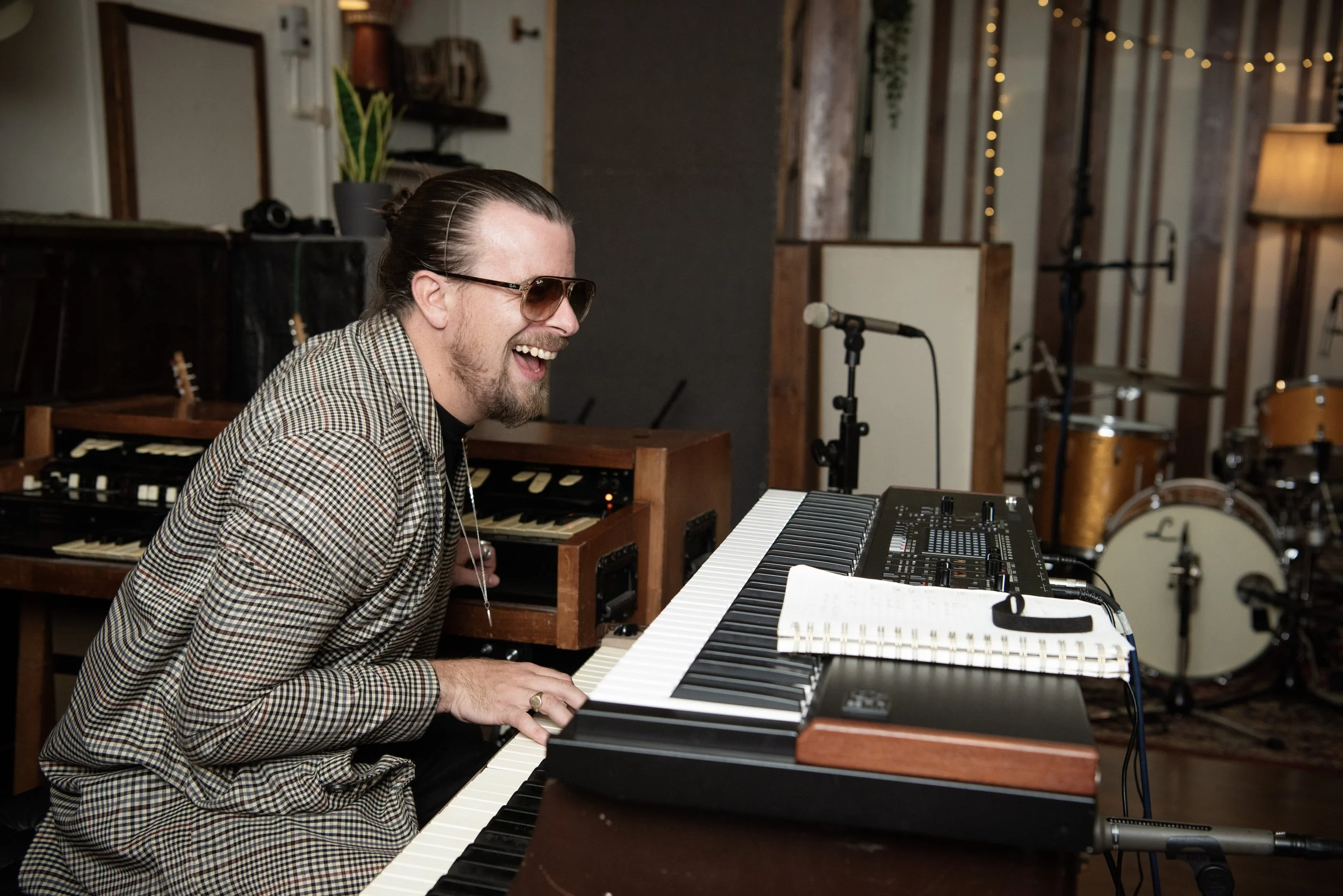 A man with glasses and a plaid shirt playing the keyboard, smiling and laughing, in a music studio with drums and microphones in the background.