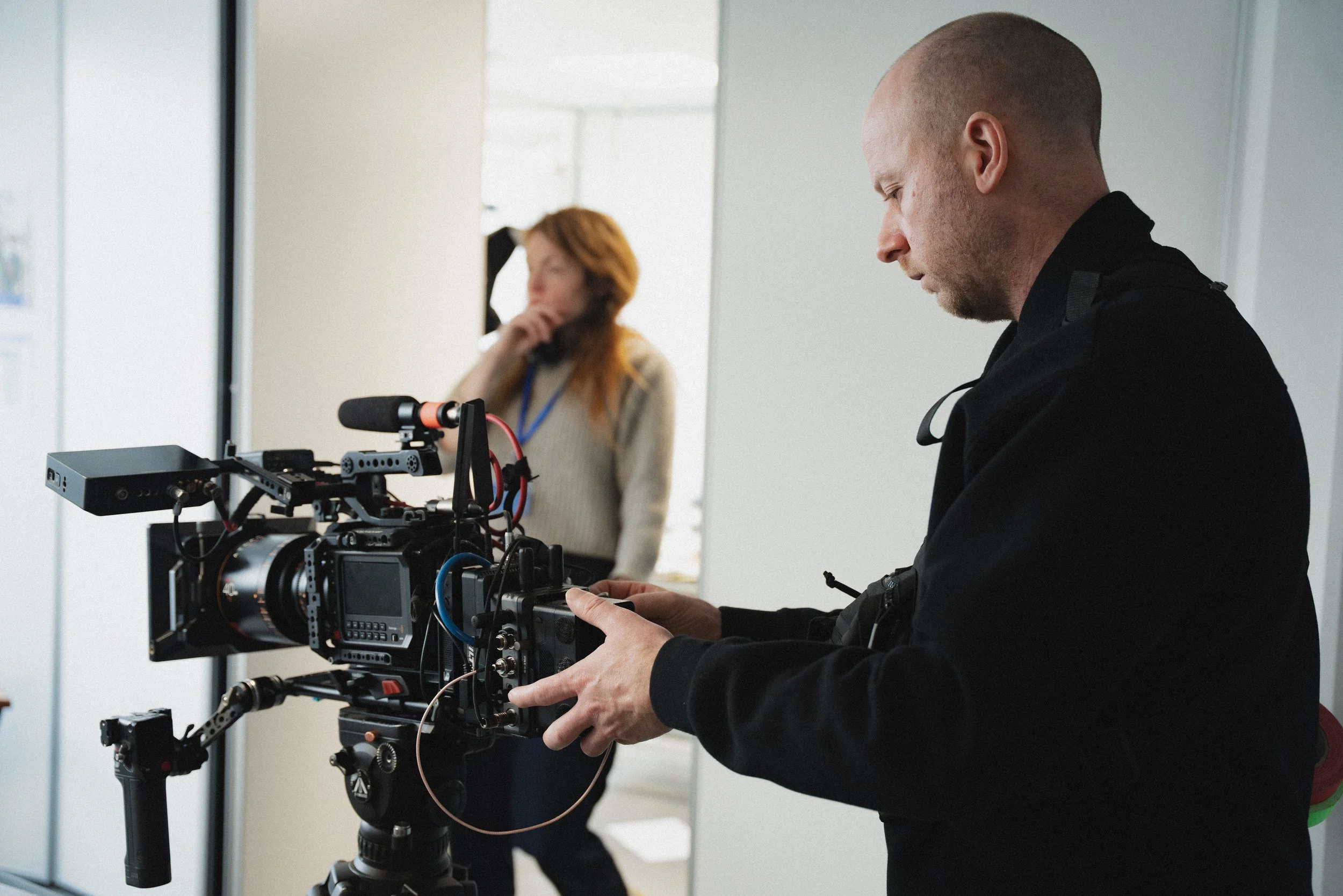A man operating a professional video camera on a tripod, with a woman in the background seemingly on a phone in an indoor space.