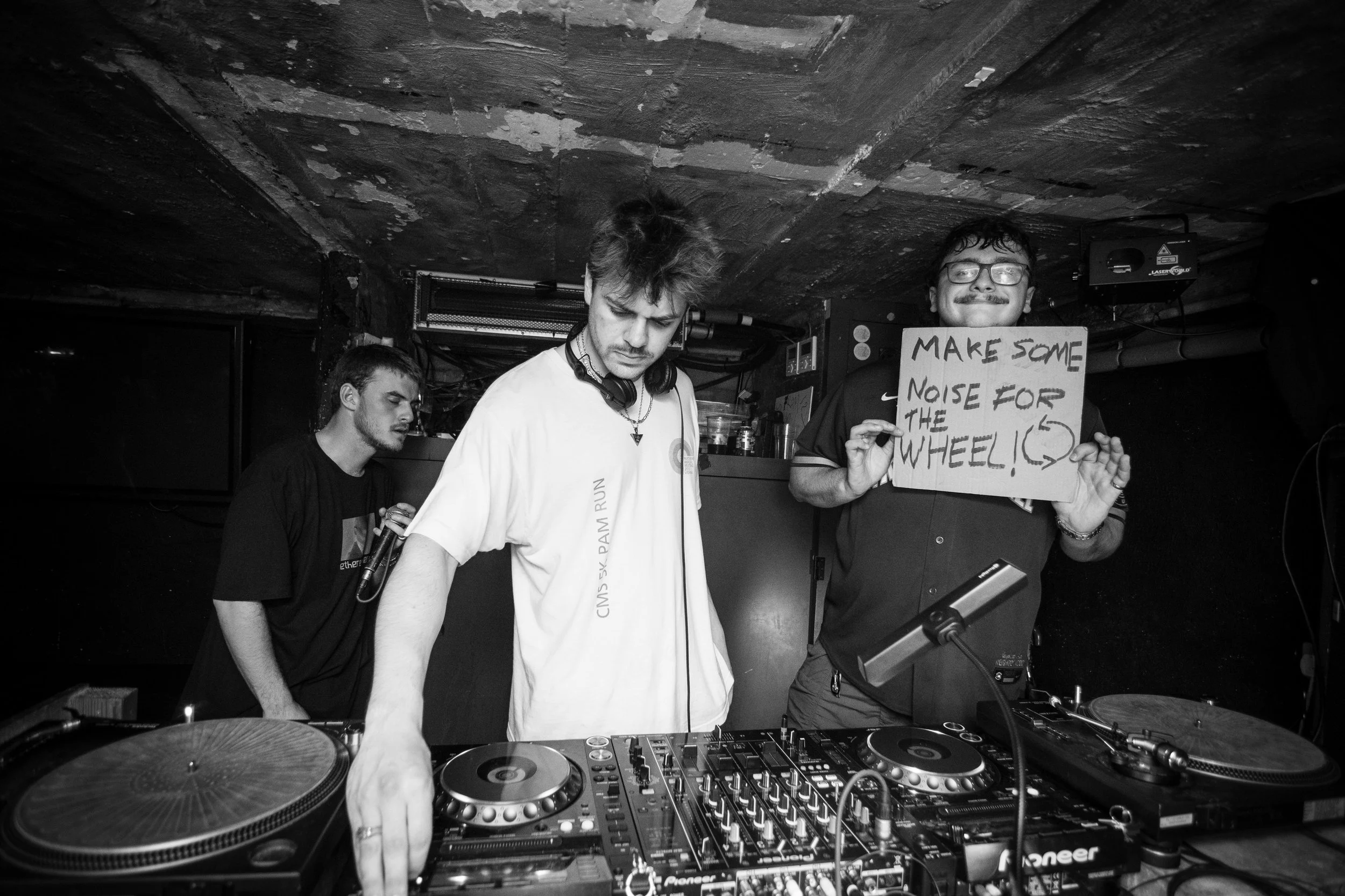 Three young men are at a DJ booth in a dimly lit club. One of them is holding a sign that reads, 'Make some noise for the wheel!' while smiling.