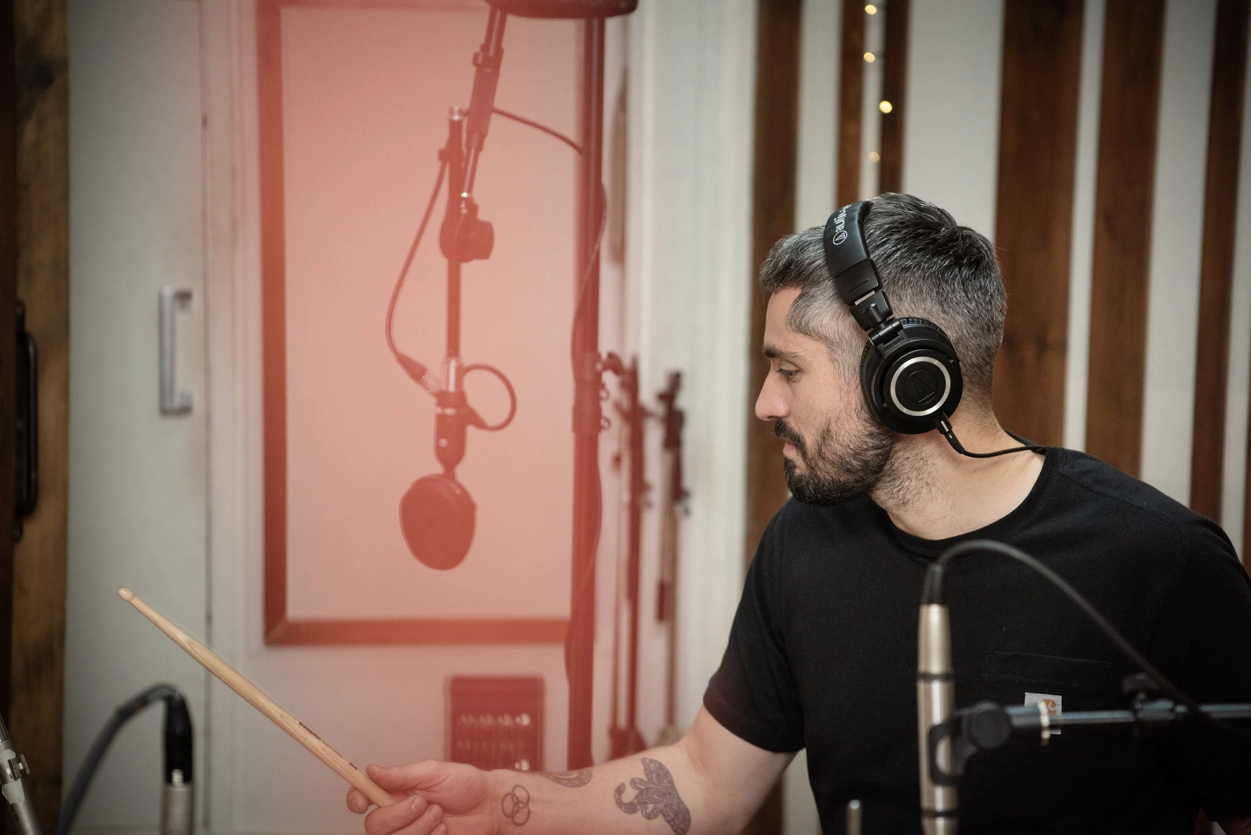 A man with tattoos on his arm, wearing headphones, is holding a drumstick and appears to be recording in a studio; a microphone and soundproofing panels are visible.