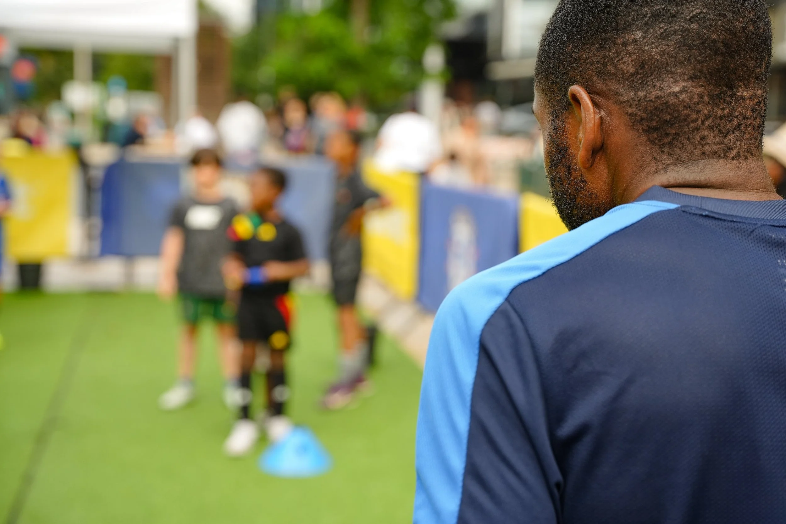 A man in a blue sports shirt observes children playing a game outside. The background shows a yellow and blue barrier, blurred figures of children, and people in a busy outdoor setting.