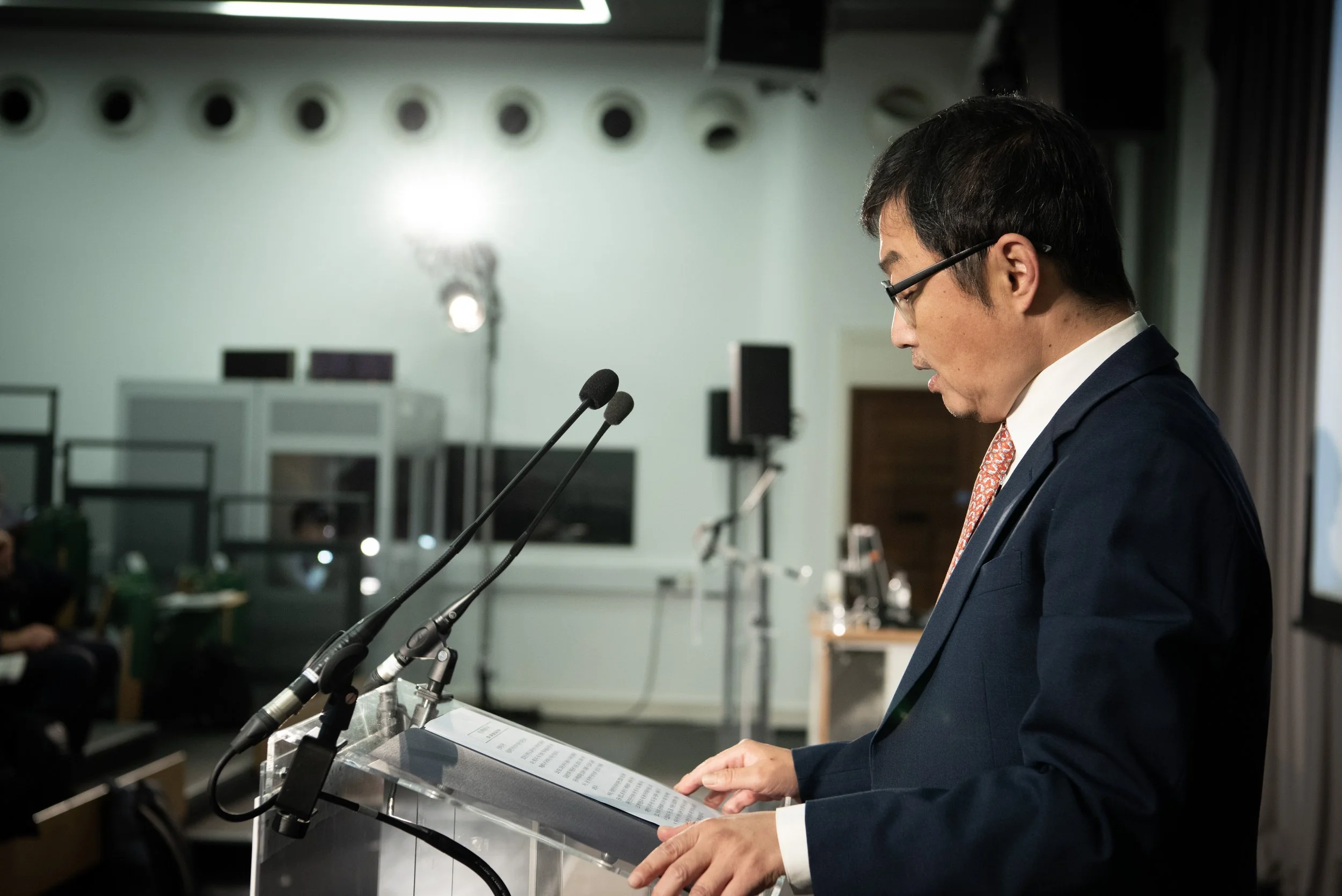 A man in a suit and glasses standing at a podium with a microphone in a conference room, reading from a document.