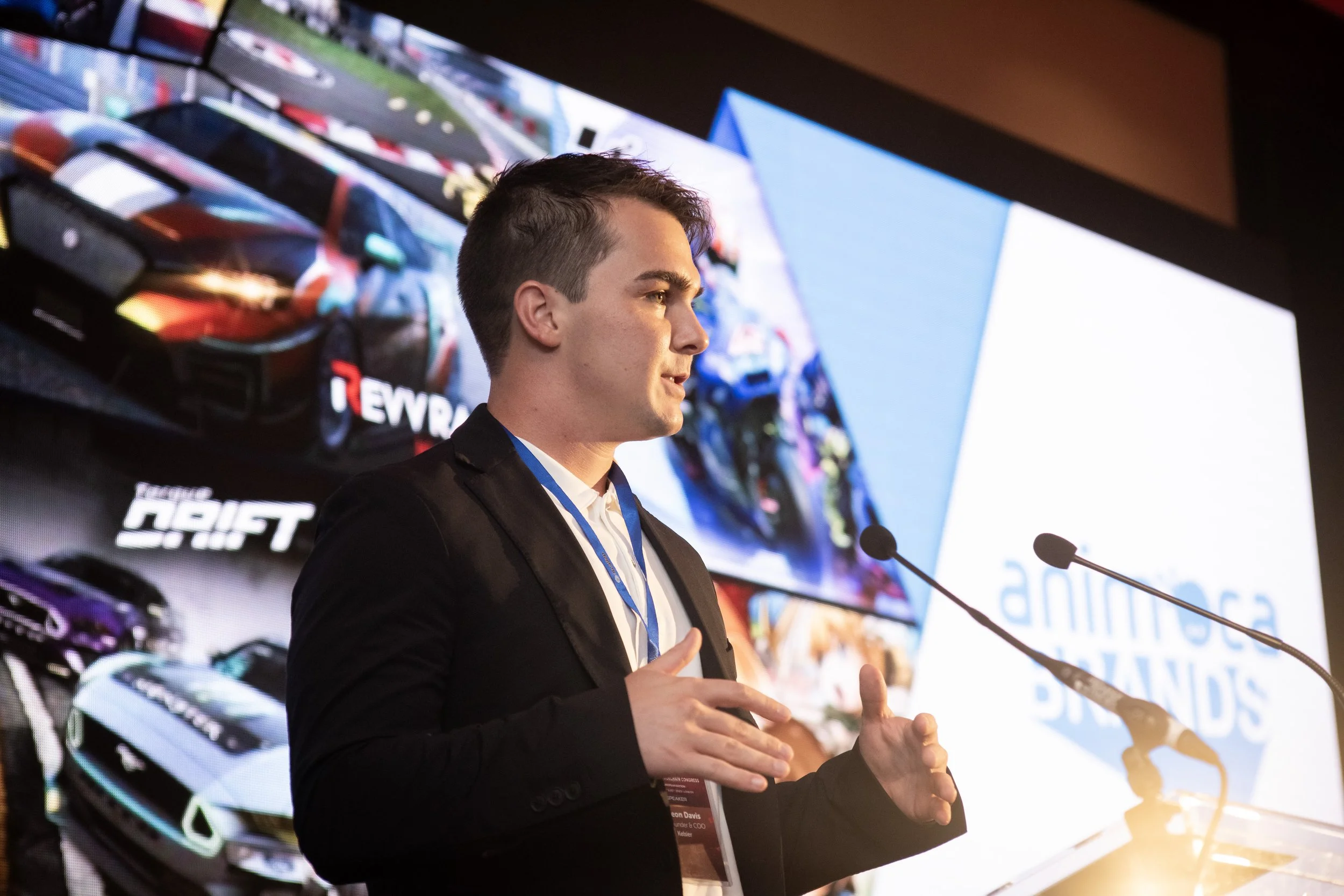 Young man in a black suit speaking at a conference with large animated screens behind him displaying car images and the Animoca Brands logo.