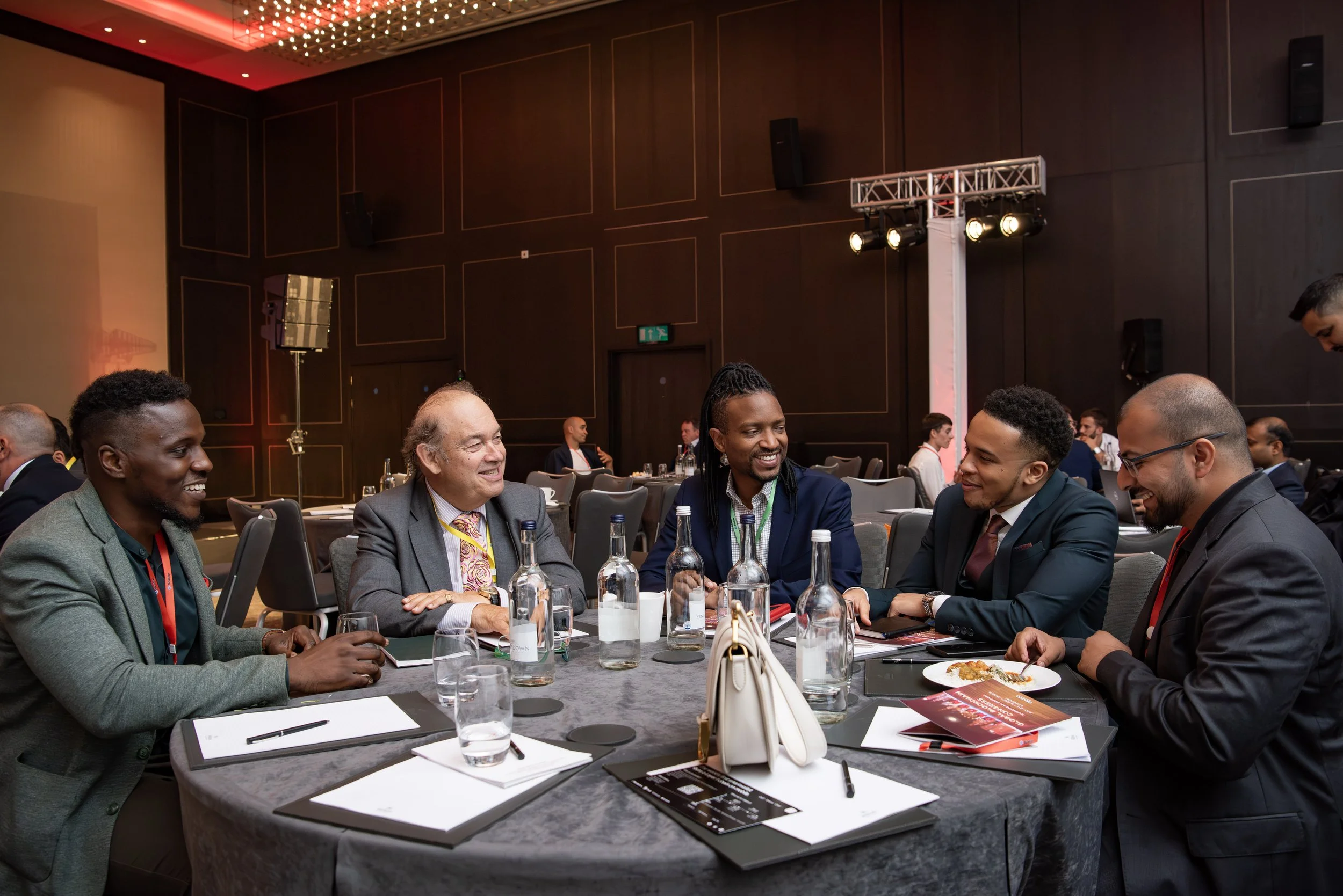 A group of five men in business attire sitting around a table at a conference or event, engaging in conversation and smiling. The table has notebooks, pens, bottles of water, and a cream-colored handbag. The background shows more people seated at tab