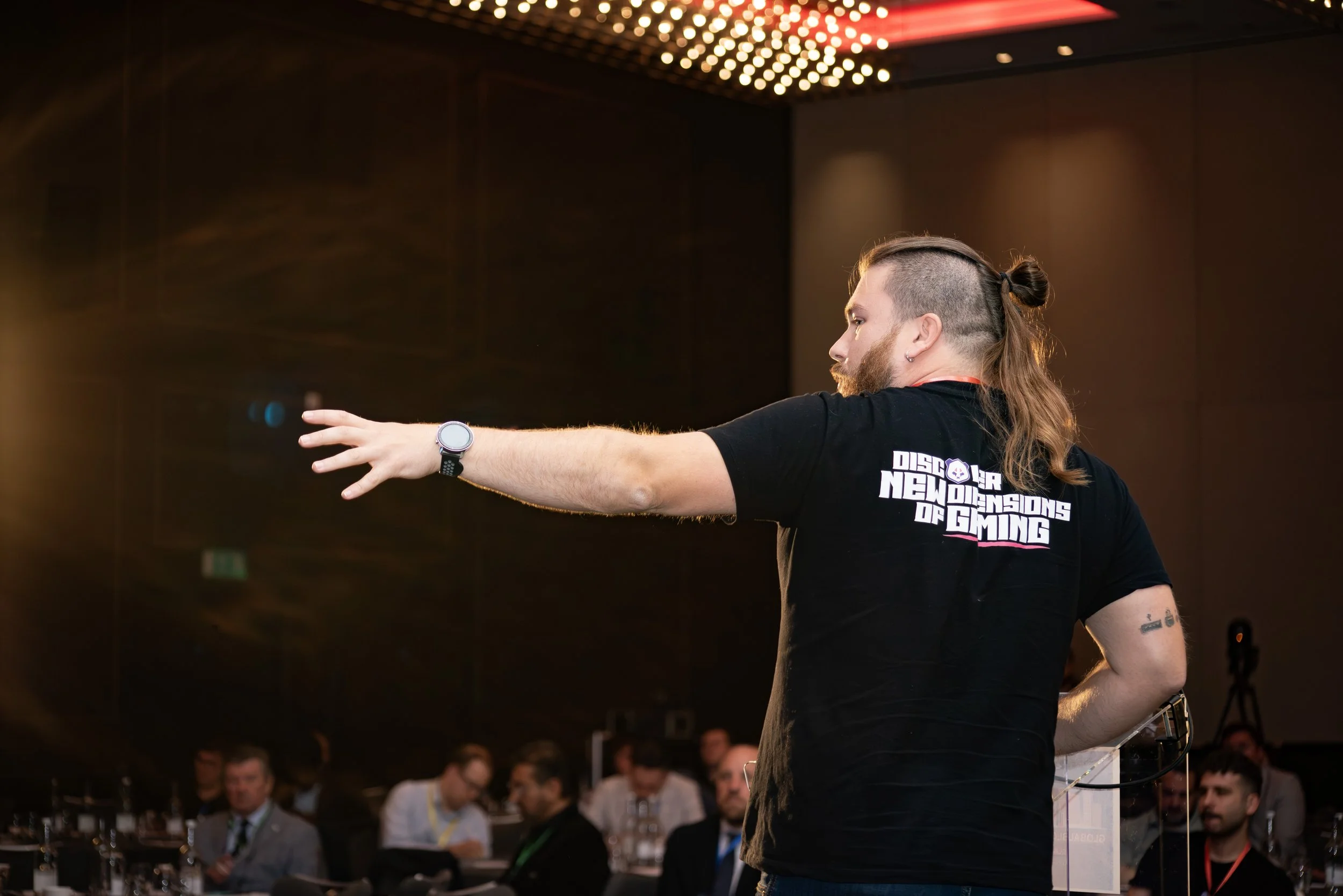 A man with long hair tied in a bun, beard, and tattoos on his left arm, wearing a black T-shirt and a watch, appears to be giving a presentation or speech at a conference or event. The T-shirt has the text 'DISCOVER NEW DIMENSIONS OF GAMING' on the b