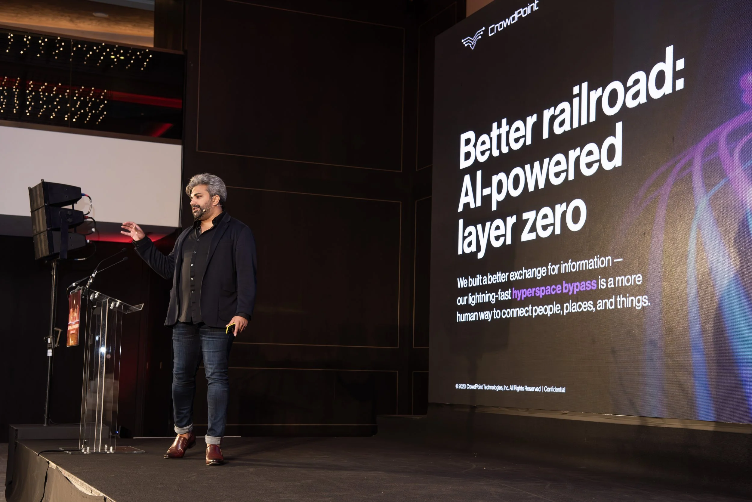A man standing on a stage giving a presentation, with a large screen behind him displaying the text "Better railroad: AI-powered layer zero" and a description about building a better exchange for information using hyperspace bypass.