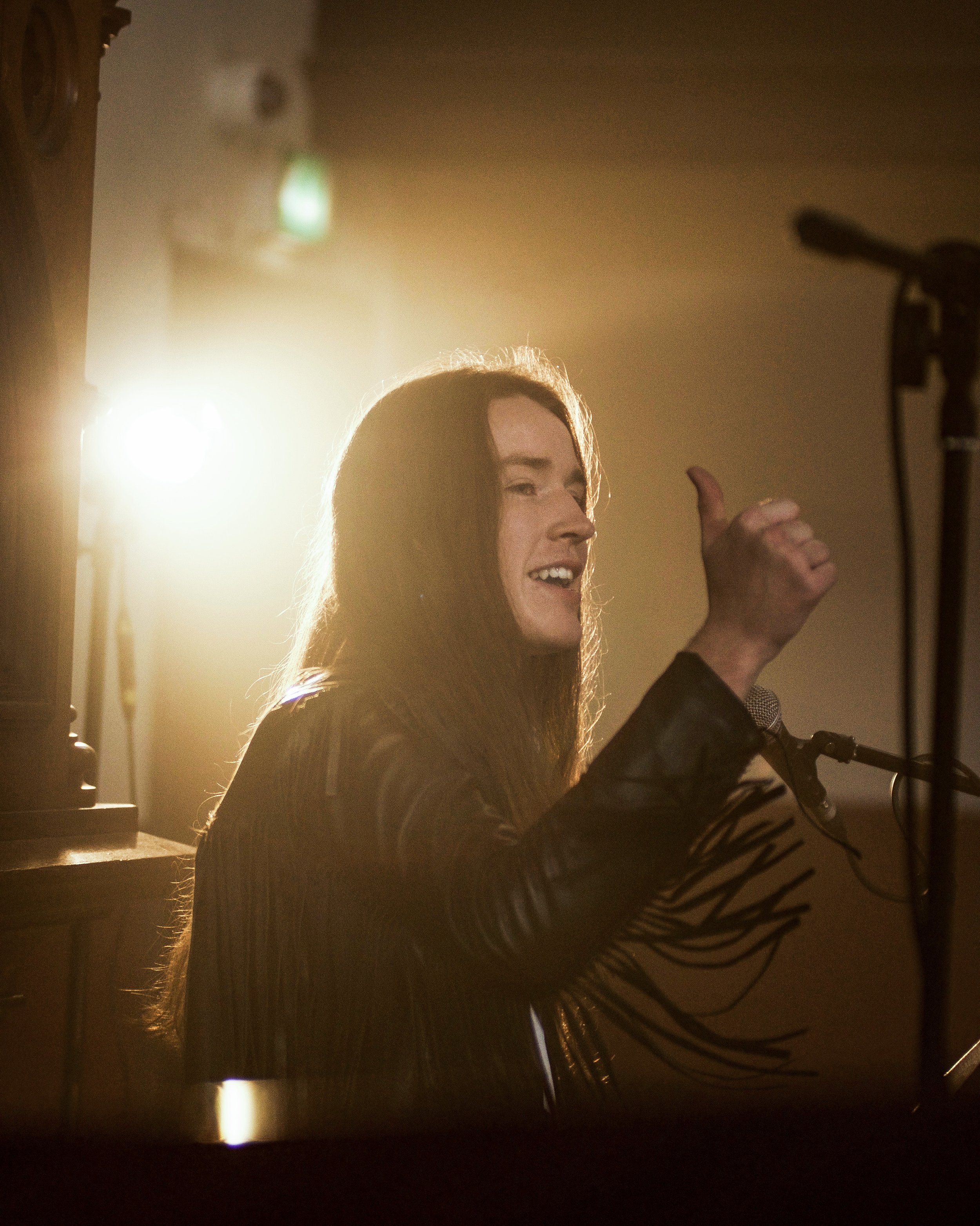A woman with long brown hair giving a thumbs-up, speaking into a microphone, with a bright backlight creating a silhouette effect.