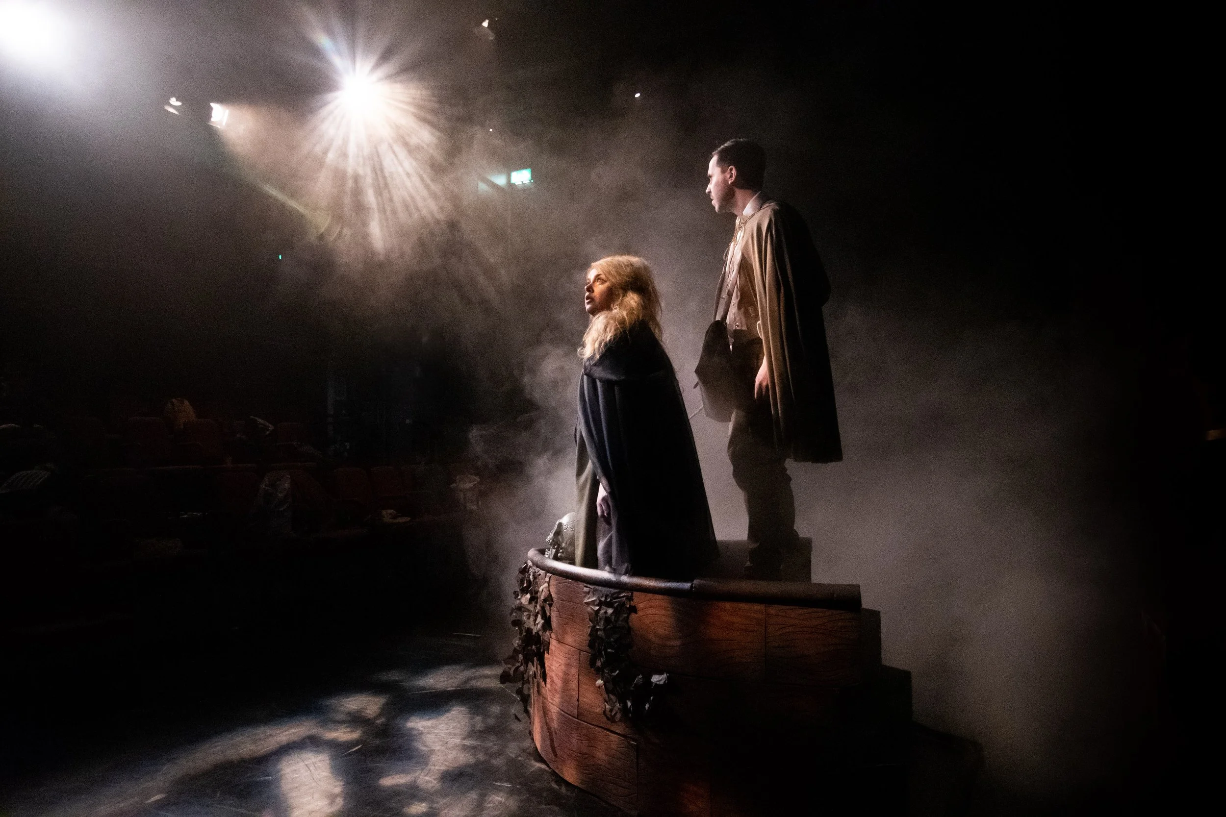Two actors on stage, facing each other, with stage lighting creating a dramatic effect, standing on a wooden platform with fog around them, in a theater setting.