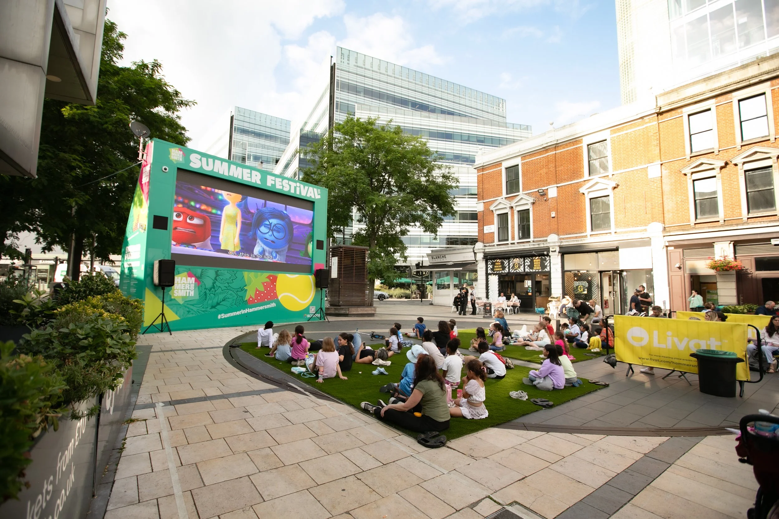People watching an outdoor movie on a large screen at a summer festival in an urban area, with some sitting on mats and others standing, surrounded by modern and historic buildings.