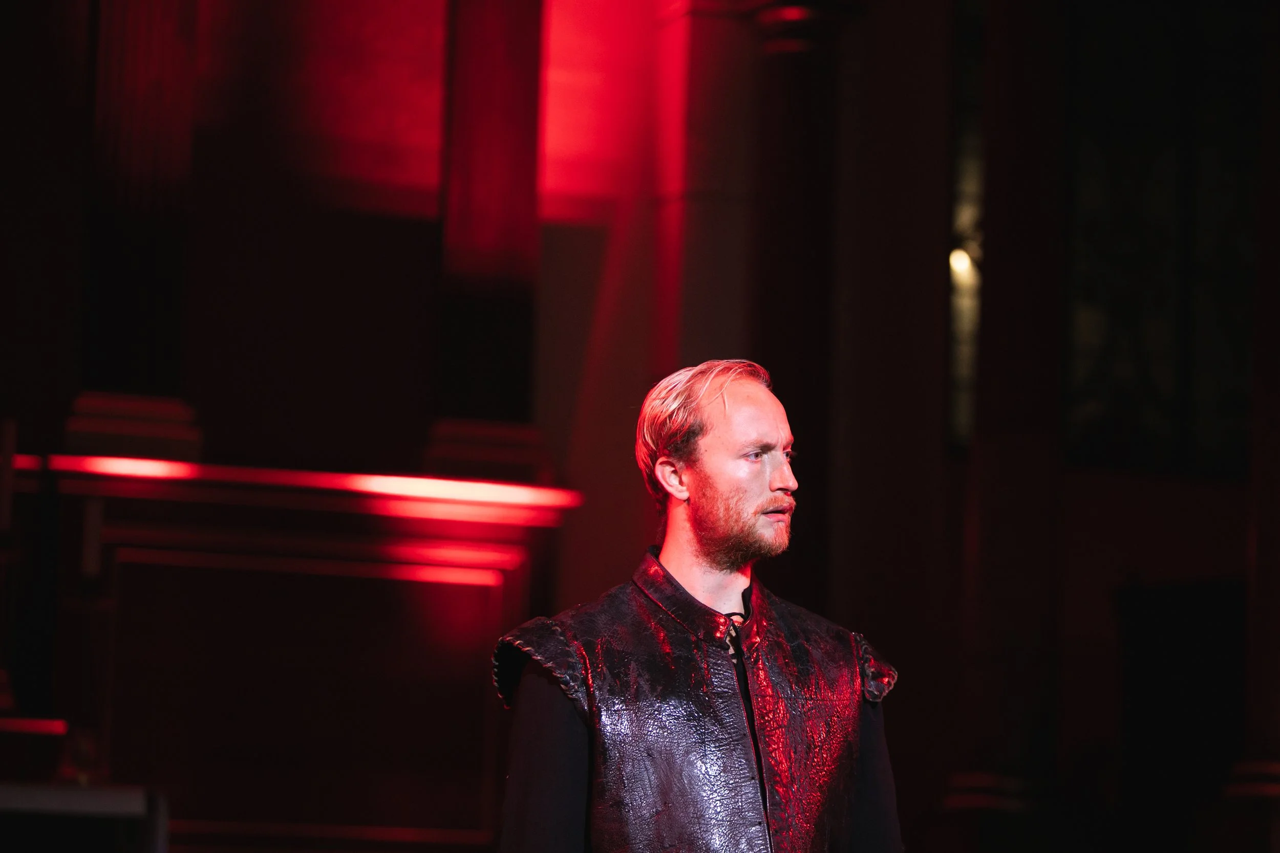 A man with blond hair and a beard is standing in a dimly lit room illuminated by red light, wearing a black vest over a black outfit, with a serious expression on his face.