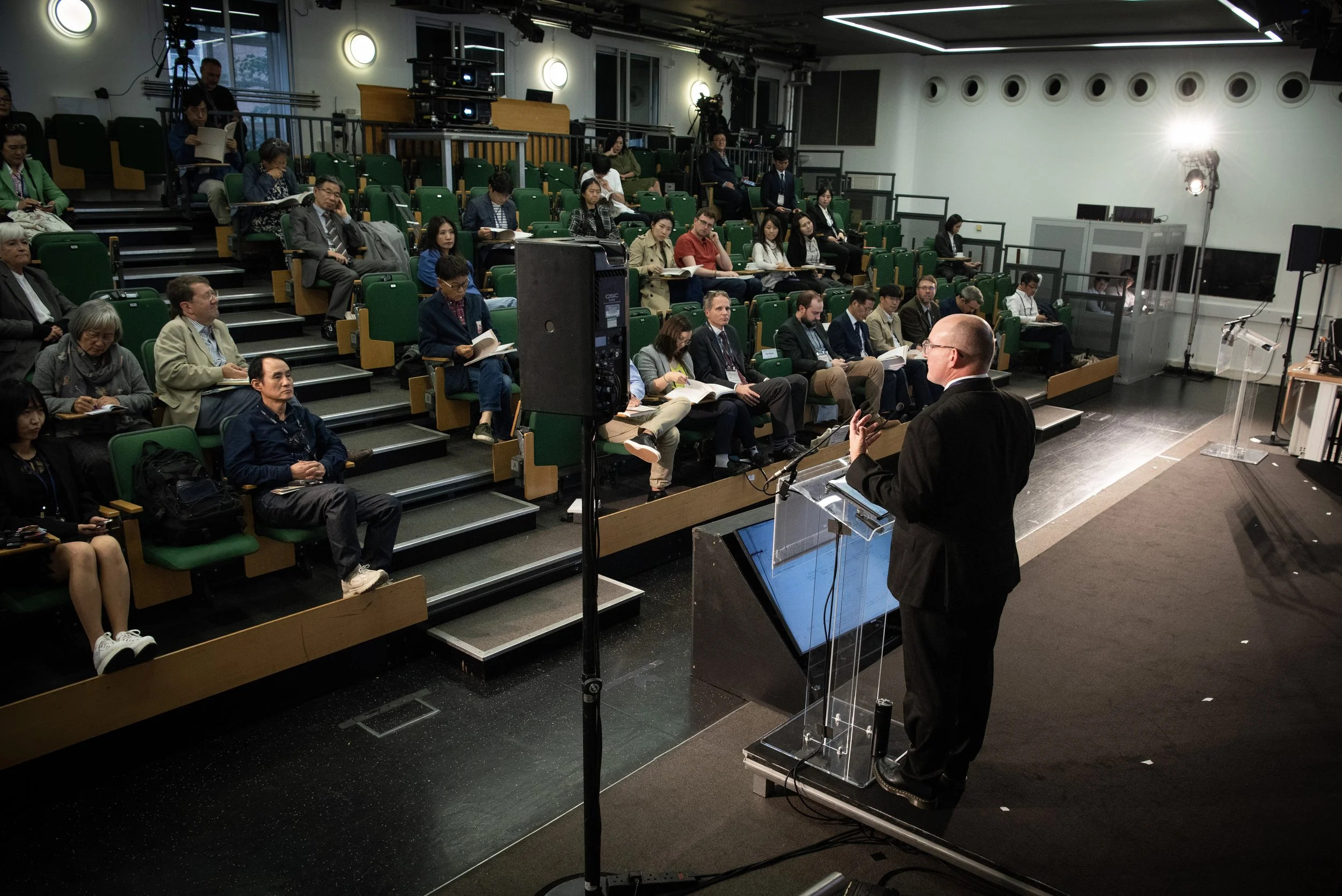 A man in a black suit giving a presentation to an audience seated in green chairs in a lecture hall.