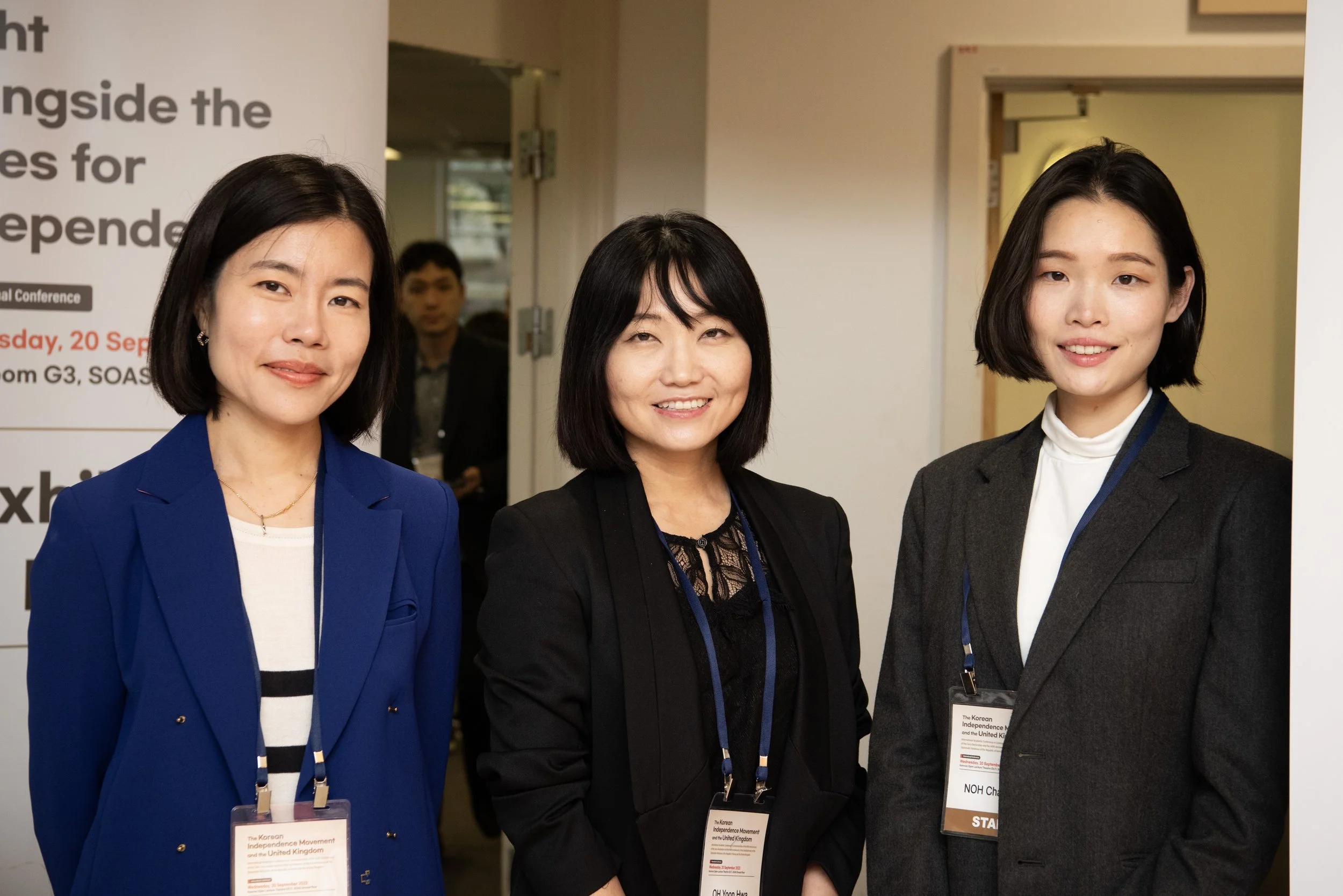 Three women smiling at an indoor conference, wearing professional attire with name badges around their necks.