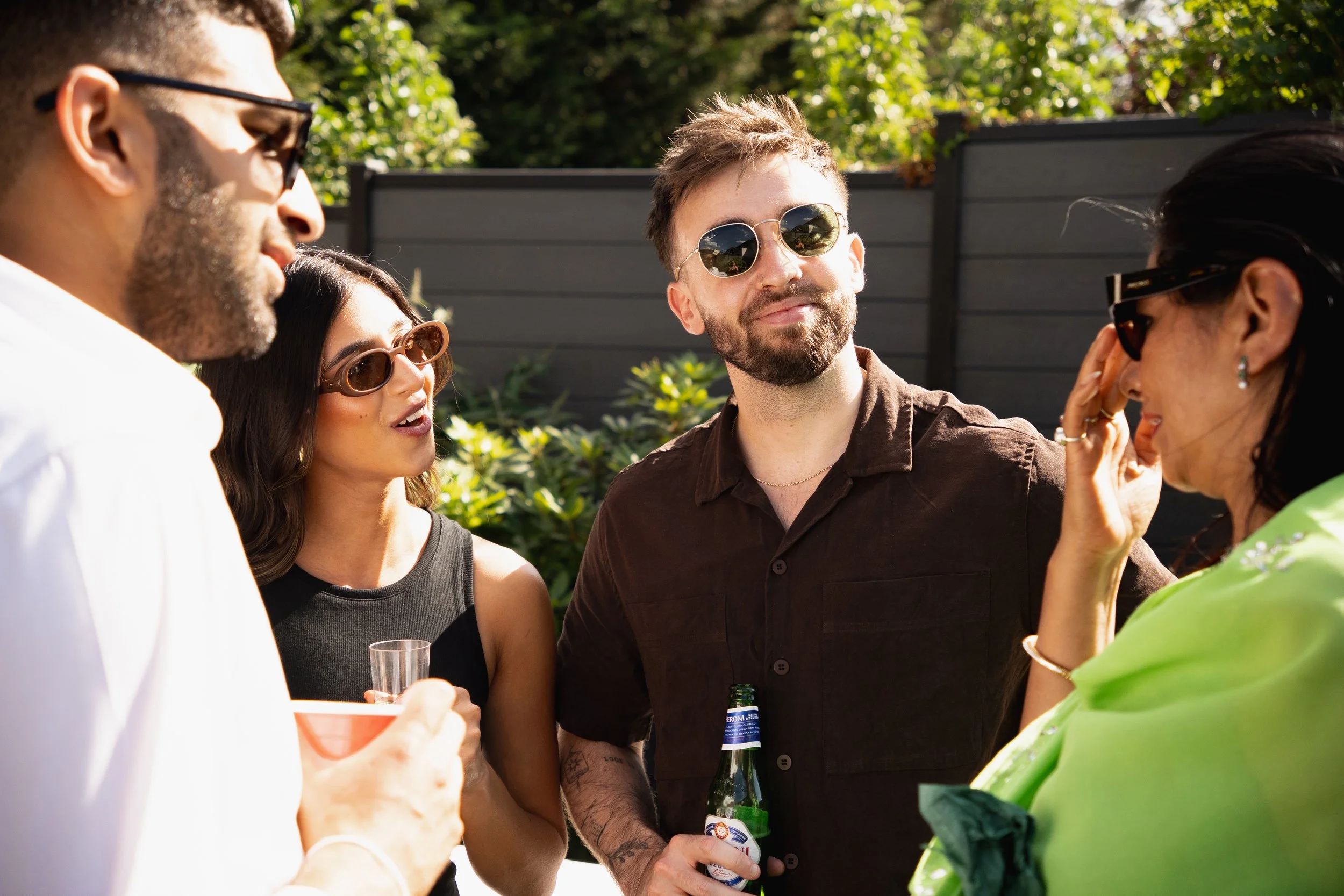 Group of five friends talking outdoors on a sunny day, holding drinks, wearing sunglasses, and smiling.