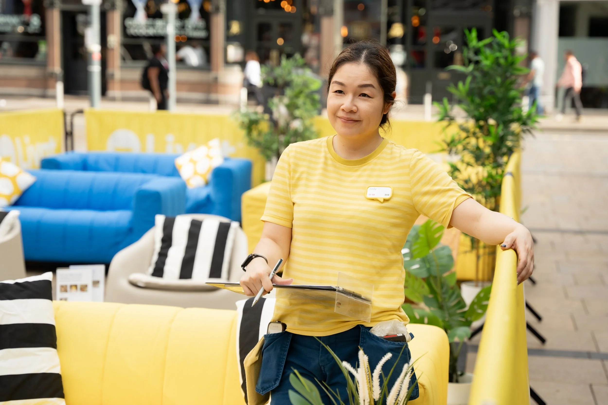 A woman in a yellow striped shirt standing outside in a furniture store or outdoor mall, holding a clipboard and pen, with colorful couches and greenery in the background.