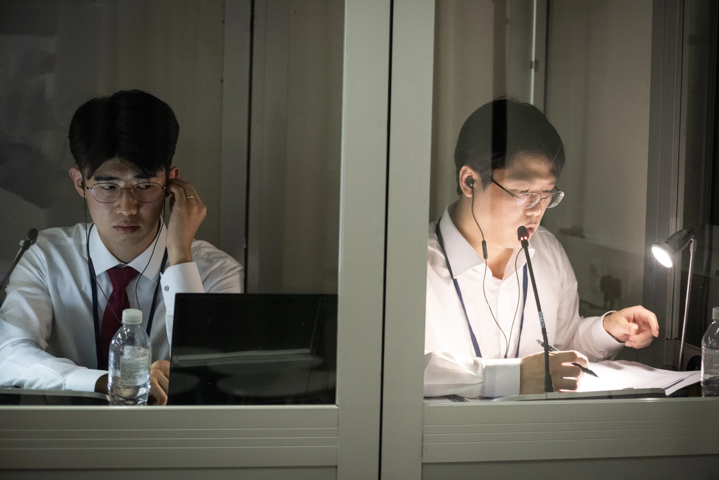 Two men wearing white shirts and glasses, working at their desks separated by glass panels in an office. The man on the left is adjusting his earphones, and the man on the right is speaking into a microphone while reading documents under a desk lamp.