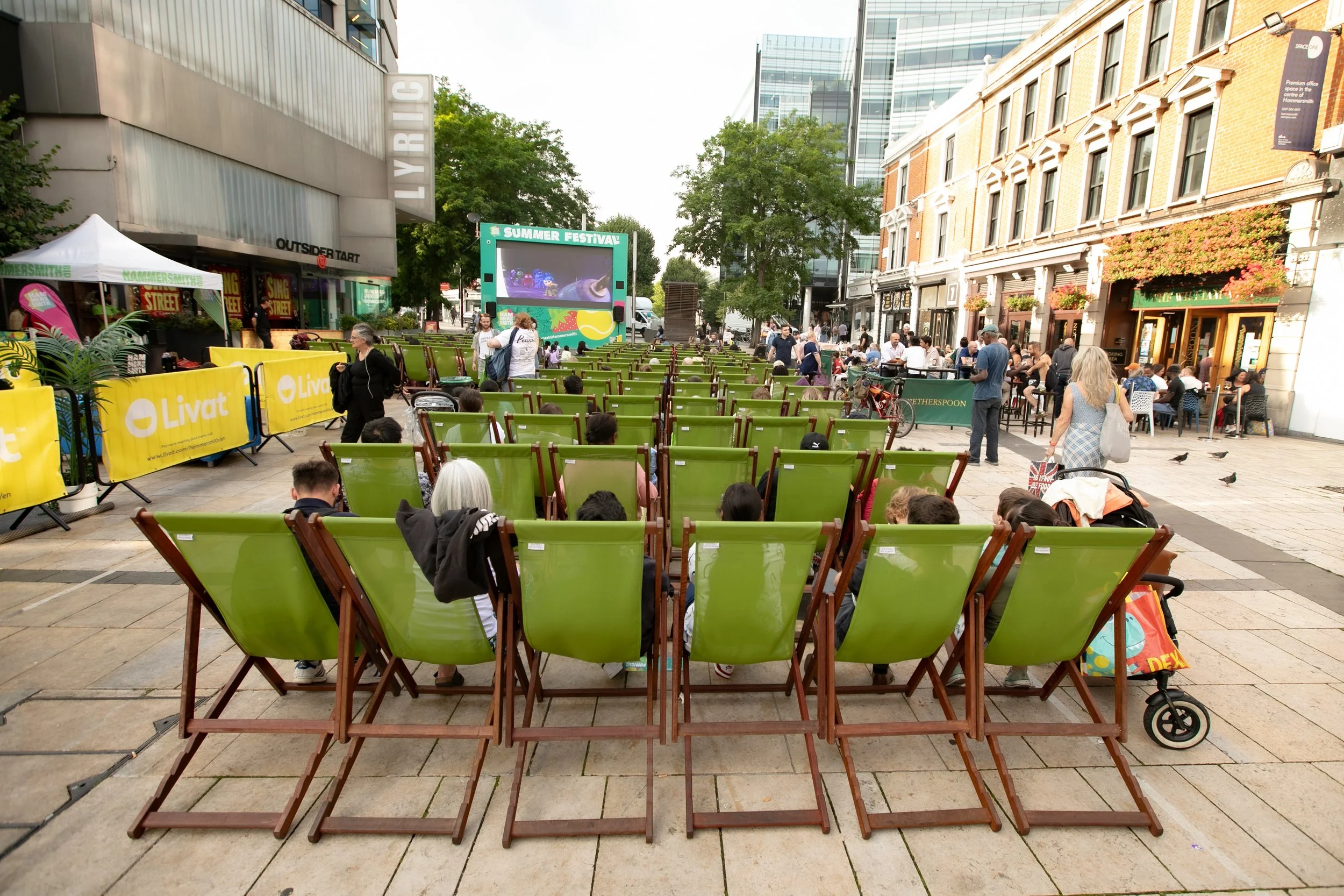 People seated in green outdoor chairs arranged in rows at an outdoor event in an urban area, with a children’s summer festival on a large screen in the background, surrounded by buildings and trees.