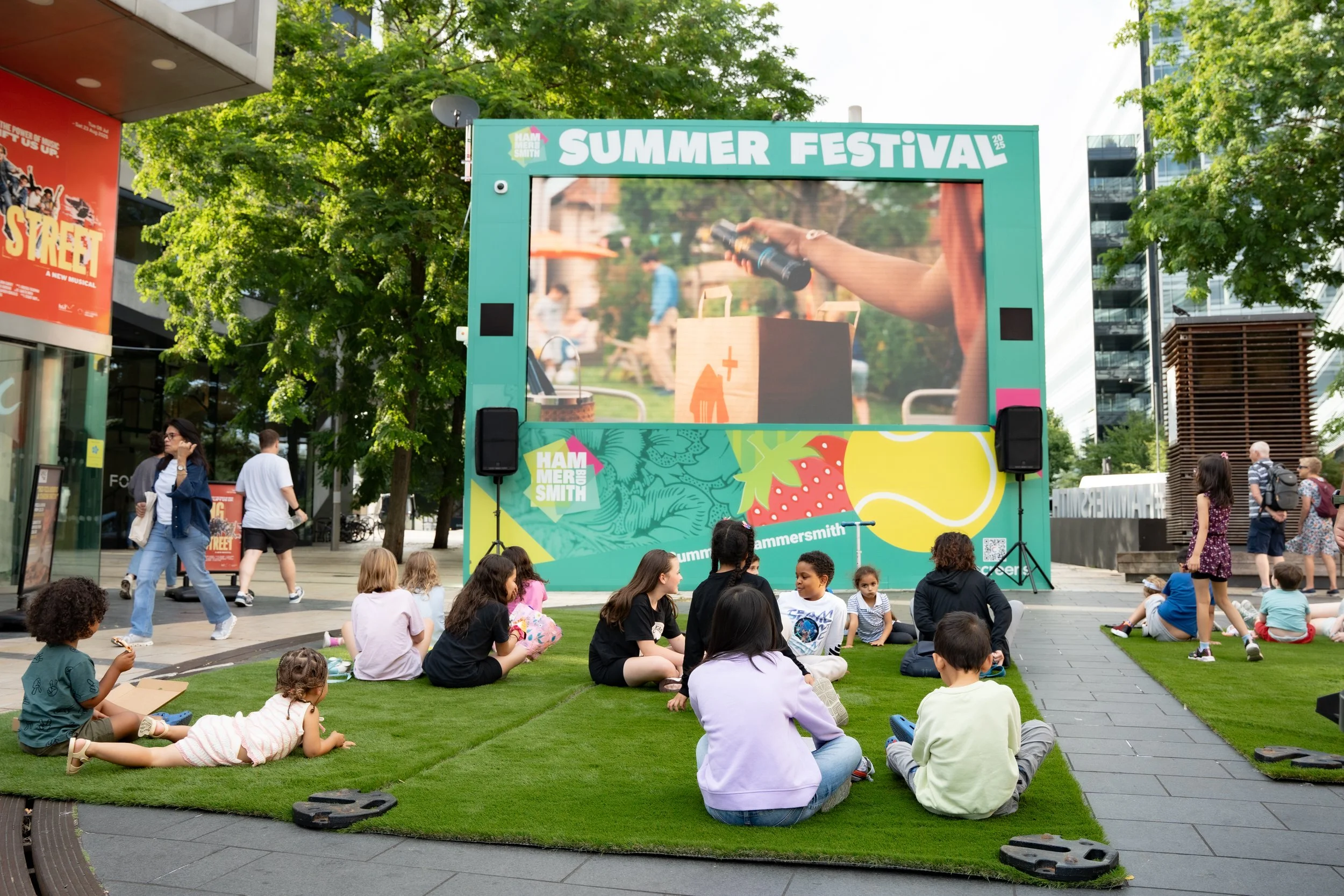 People gather outdoors in an urban plaza to watch a large outdoor screen displaying a video on the theme of a summer festival, with some sitting on green artificial grass and others walking by, surrounded by trees and buildings.
