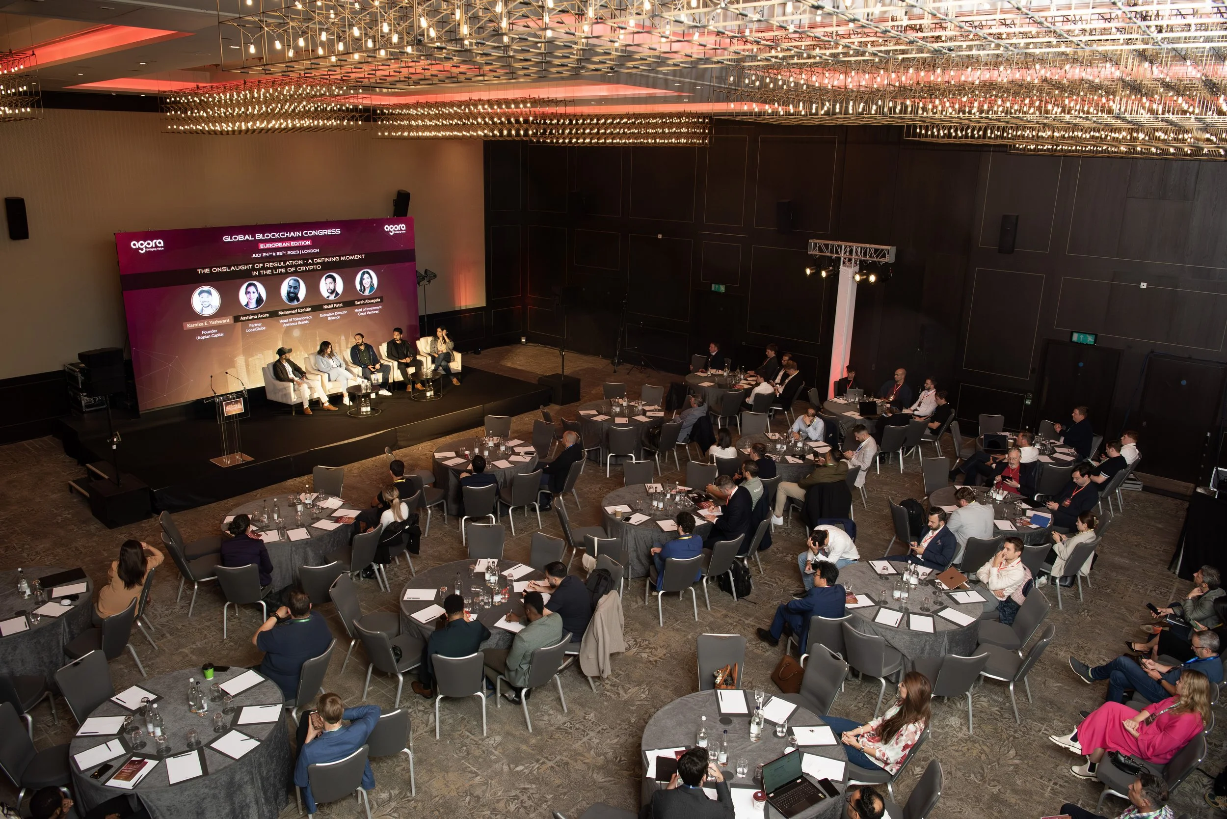 Conference room with round tables, a stage with a panel discussion, and an audience attending a blockchain event. The stage has five panelists, and a large screen behind them displays the event details.