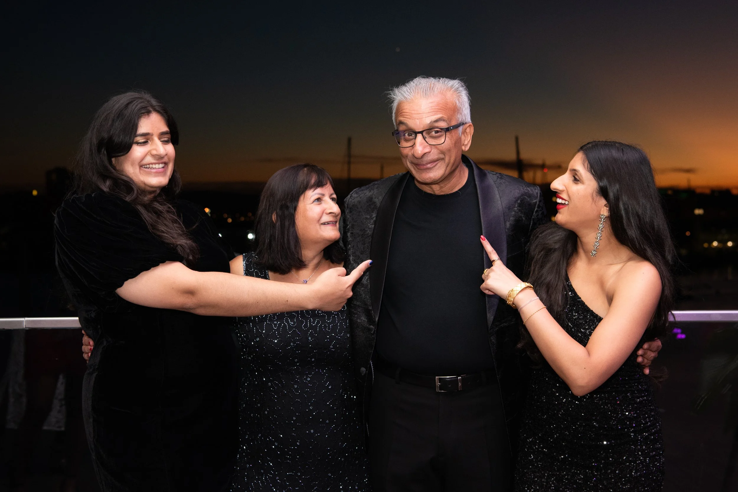 A group of four people, including one man and three women, dressed in formal attire, smiling and pointing at each other during a sunset or evening event.