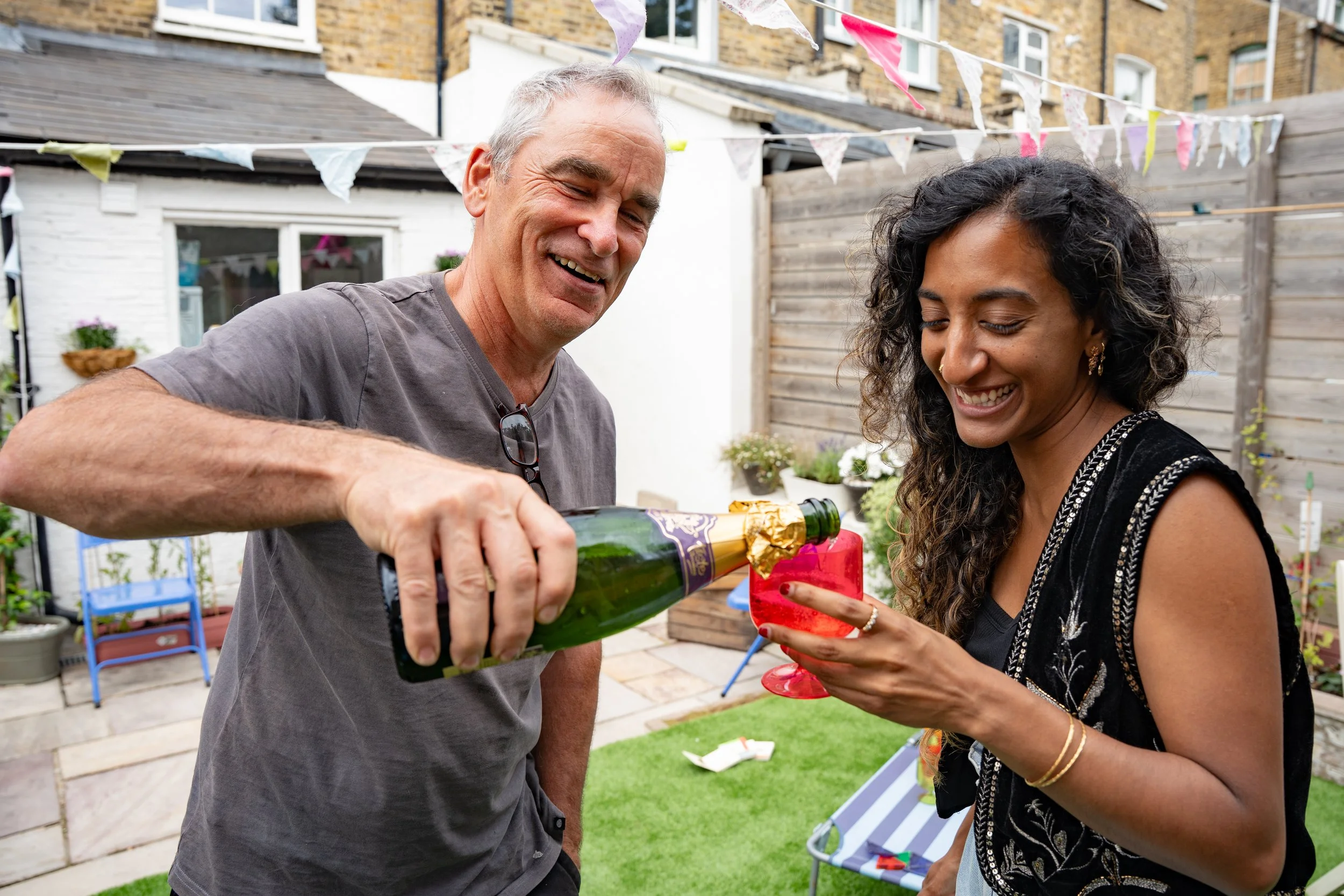A man is pouring champagne into a woman's glass during a garden celebration, with bunting and potted plants in the background.