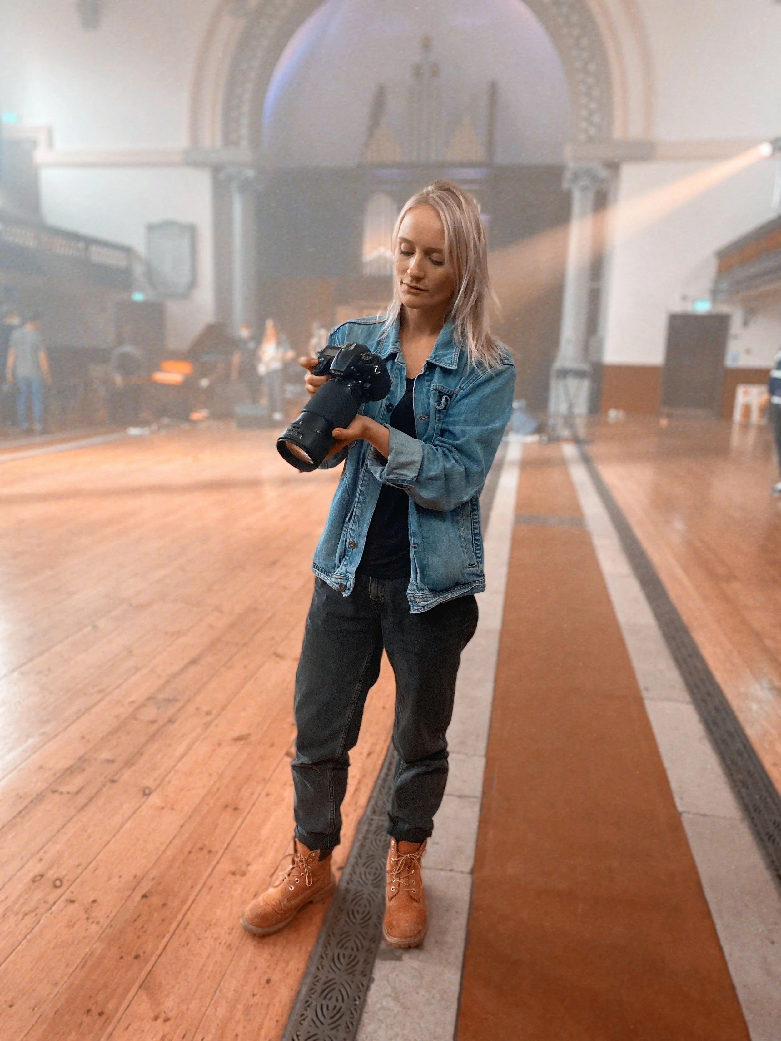 A woman with blonde hair wearing a denim jacket, black pants, and tan boots, holding a camera in an indoor venue with wooden floors, stage, and people in the background.
