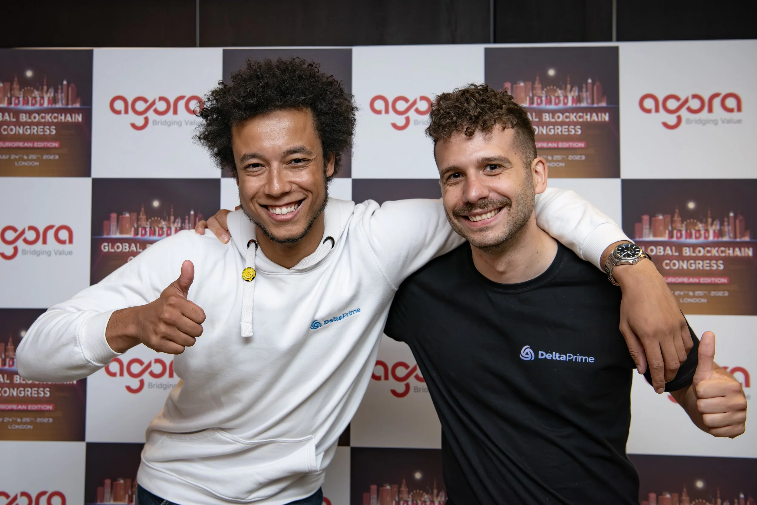 Two men smiling, with arms around each other's shoulders, posing in front of a backdrop with logos that read 'agora' and 'Global Blockchain Congress'. Both are giving thumbs-up gestures, one in a white Delta Prime hoodie and the other in a black Delt