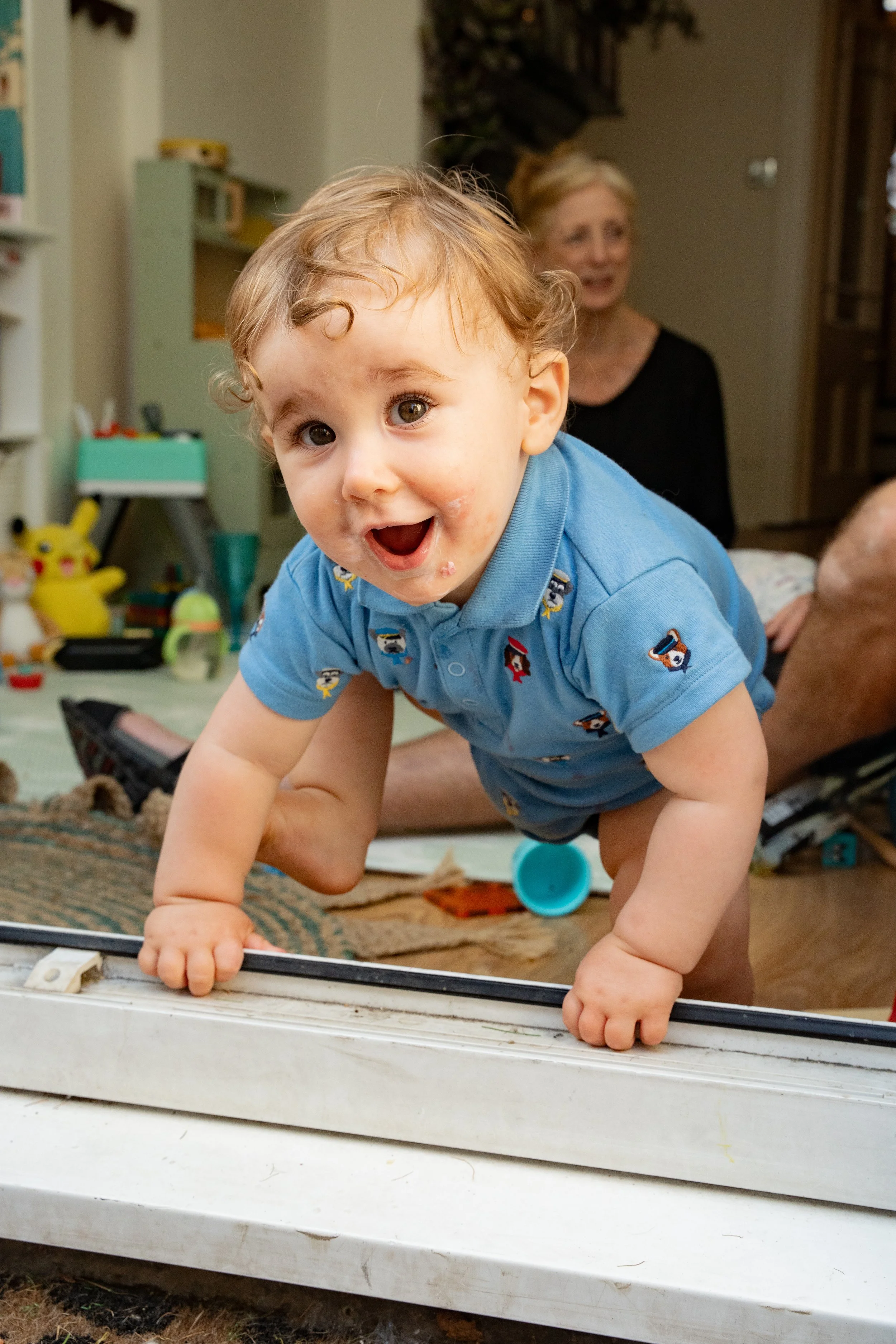 A young child crawling on a door threshold inside a home, with an older woman sitting in the background. The child is wearing a blue shirt with cartoon characters and has a happy expression.