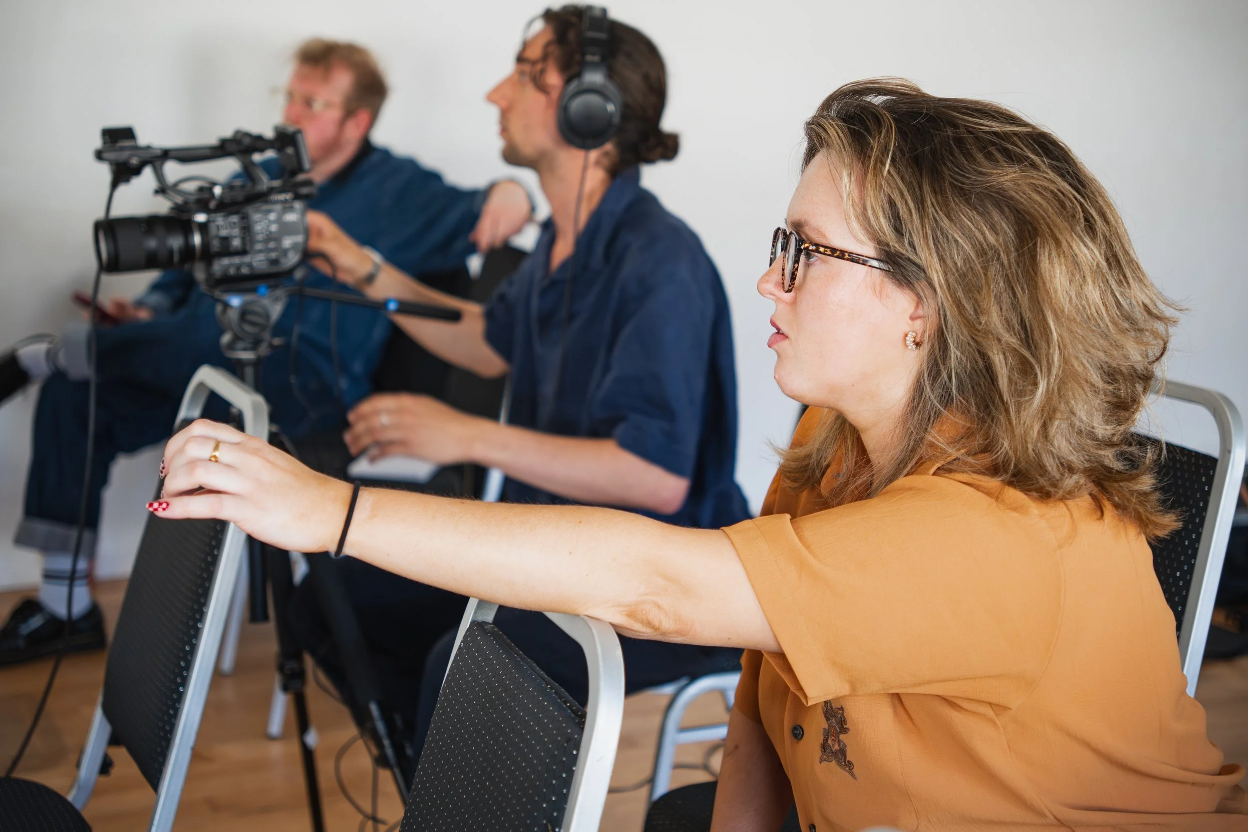 A woman with glasses and wavy hair operating a camera during a recording or interview, with two men, one with headphones, sitting behind her.