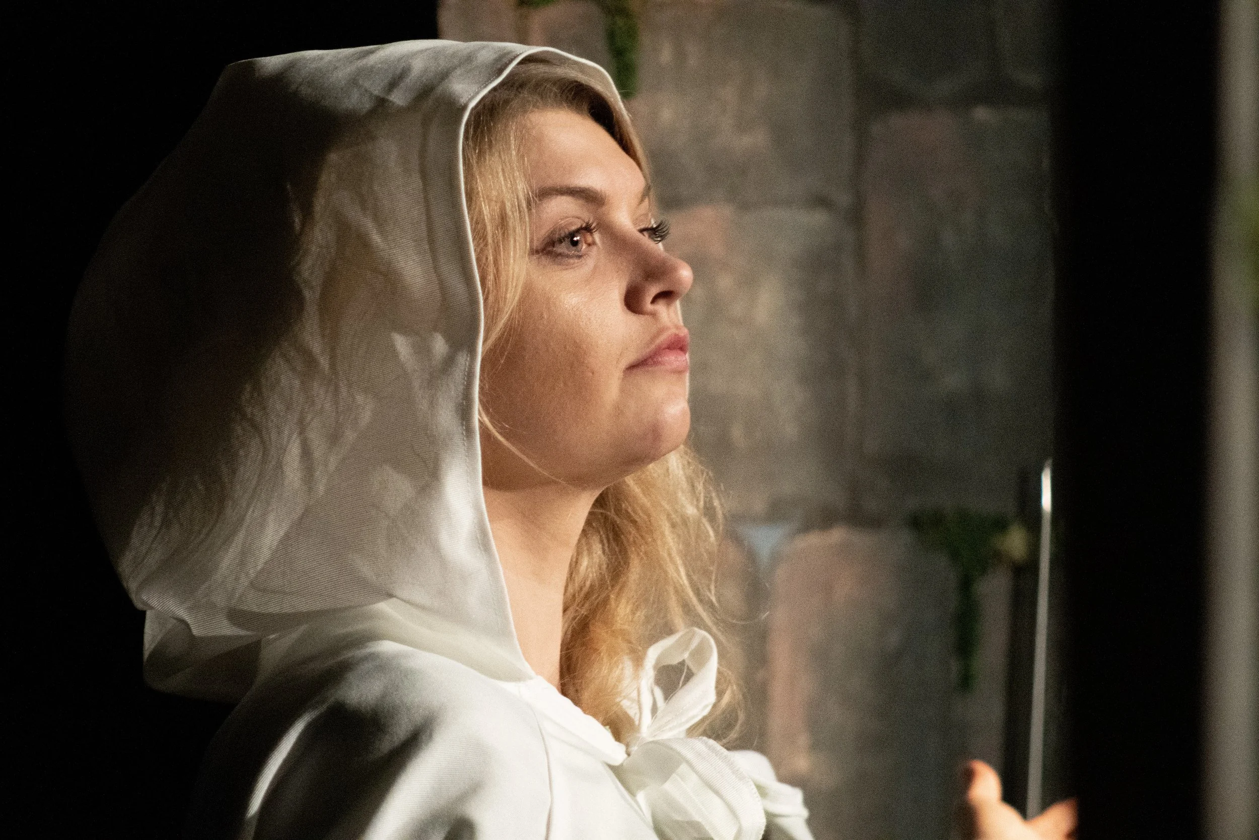 A woman with blonde hair wearing a white hooded garment, looking to the side, with a stone wall in the background.