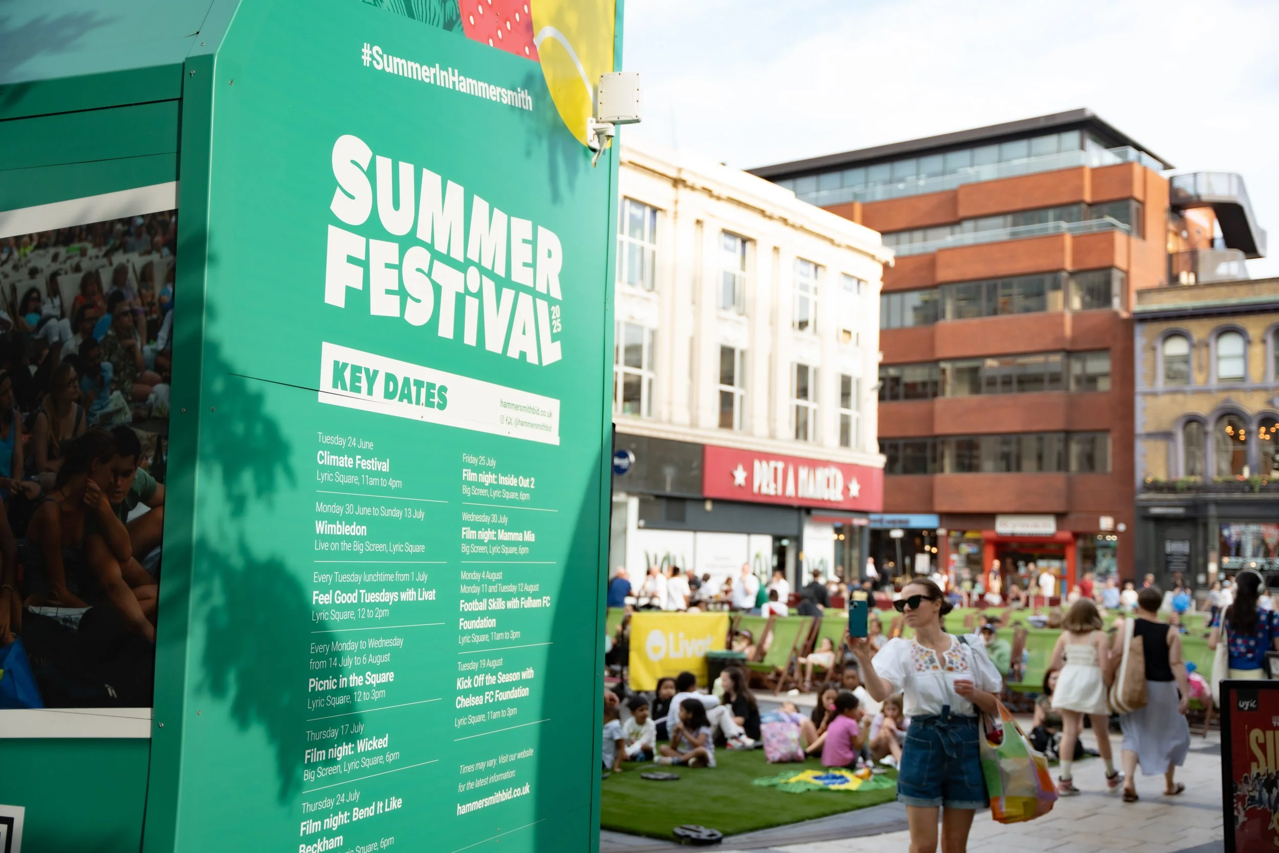 People gathered outdoors at a summer festival with a large green sign displaying event information. Some people are sitting on the grass, others are walking, and a woman is taking a photo with her phone.
