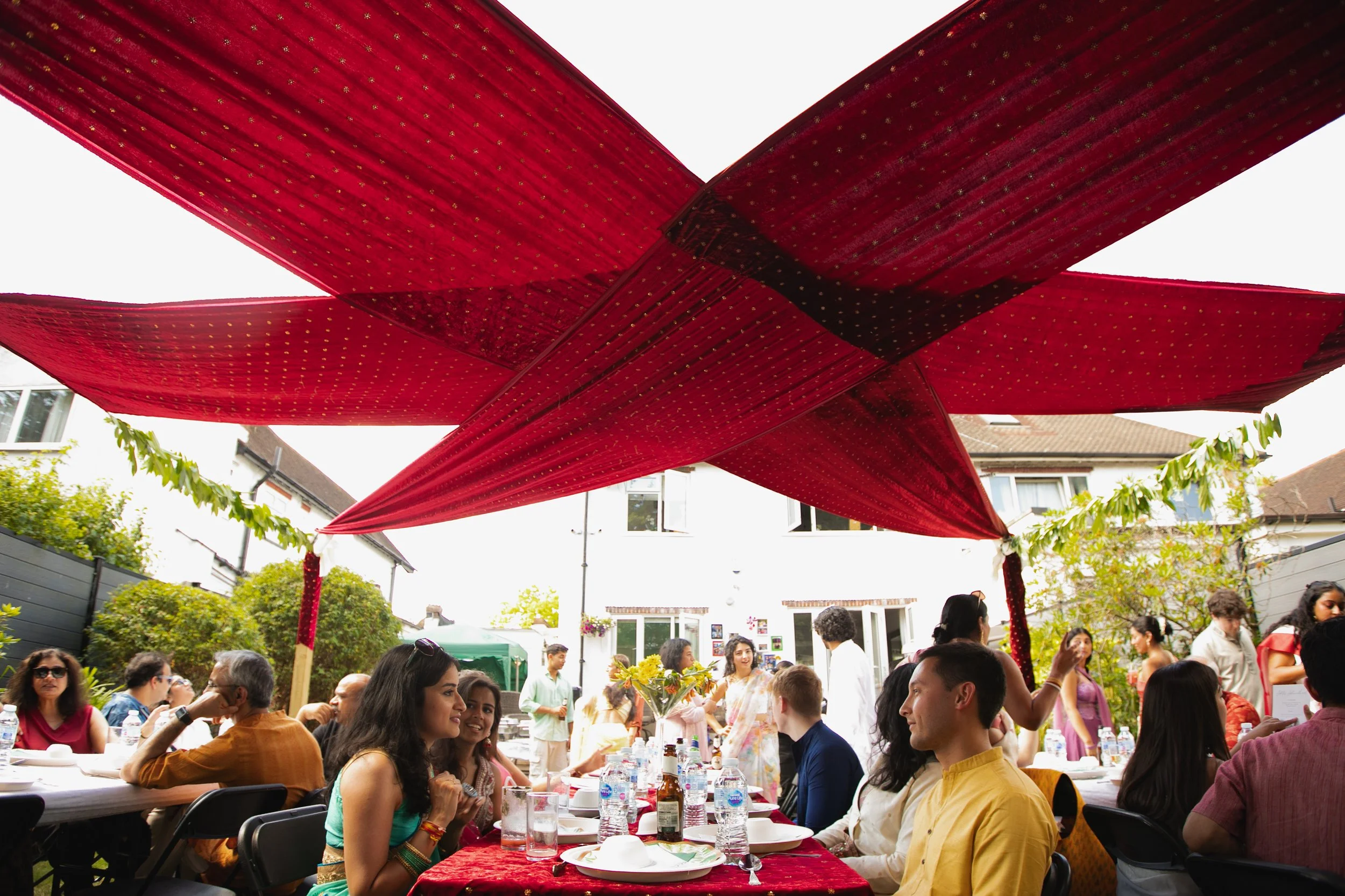 People gathered at a backyard party under a large red canopy, sitting at tables with plates and drinks, engaging in conversation.