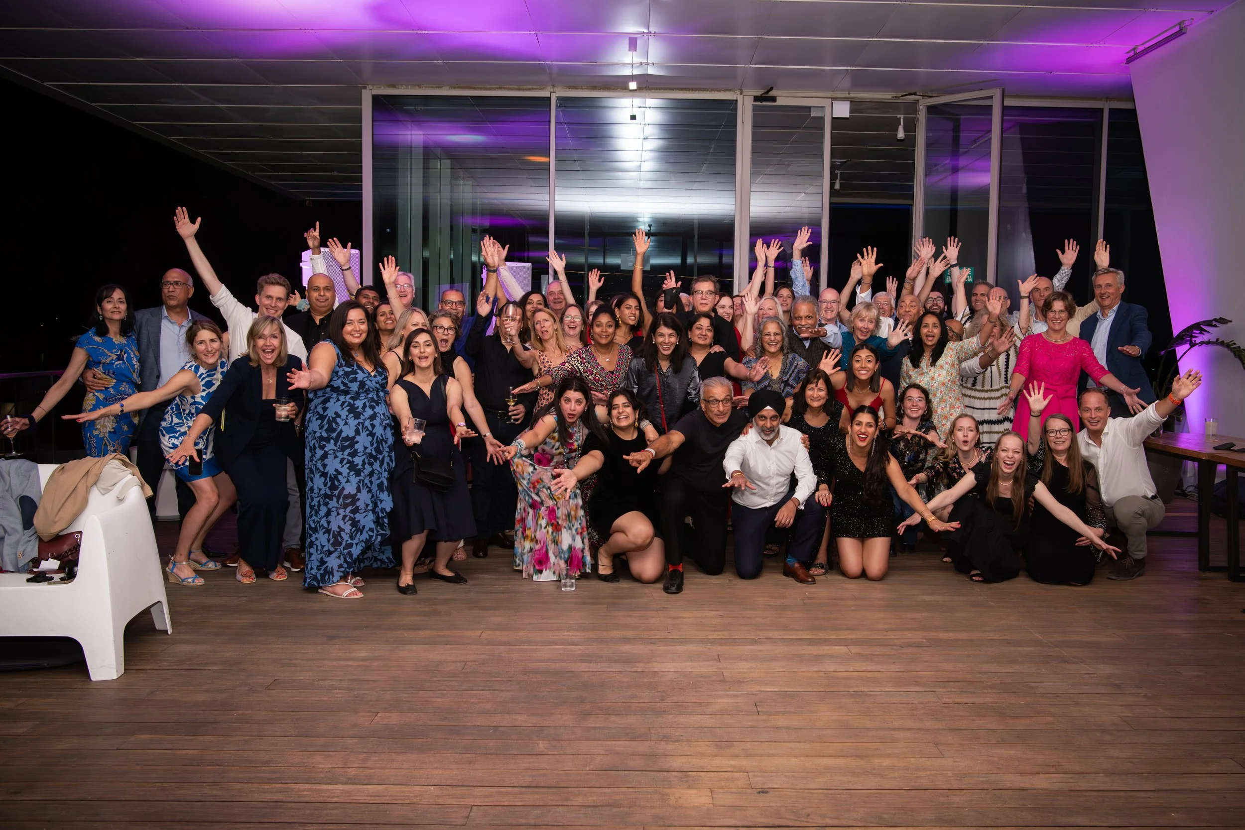 Large group of people at a celebration or party, posing together with many raising their hands, indoors with purple lighting.
