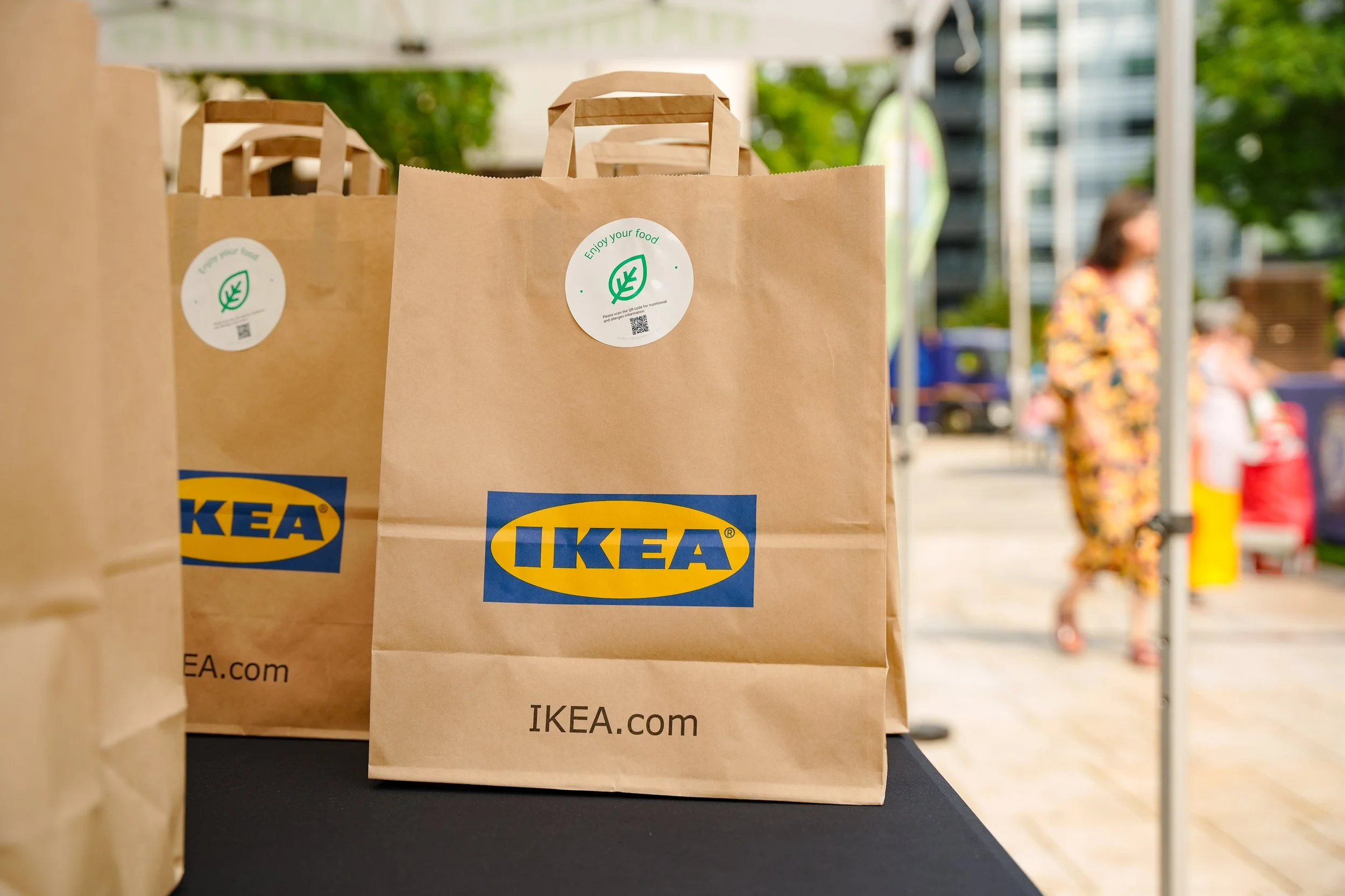 Two brown paper IKEA shopping bags with handles on a black table outdoors, with a blurred background of people and trees.