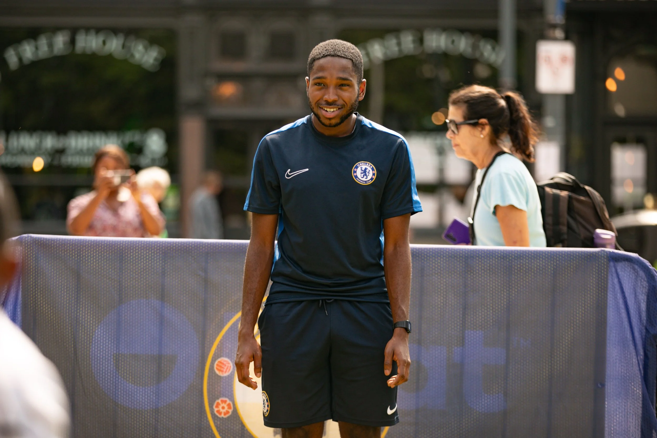 A young man wearing a Chelsea football club shirt and shorts, standing outdoors behind a table or barrier, smiling. There are people in the background, some taking photos.