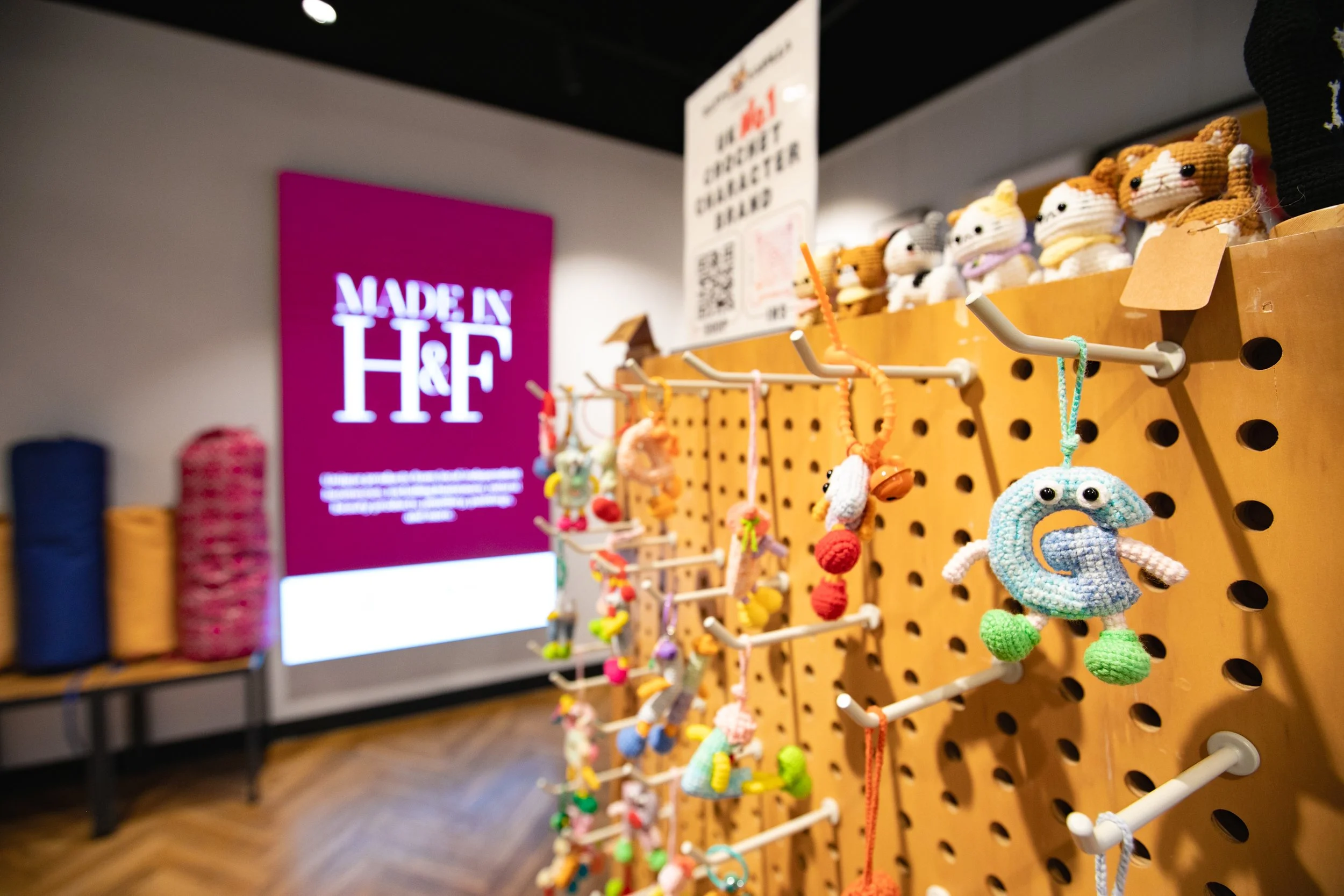 Colorful handmade animal-themed plush toys hanging on a perforated wooden display board at a store, with a pink "Made in H&F" sign in the background.